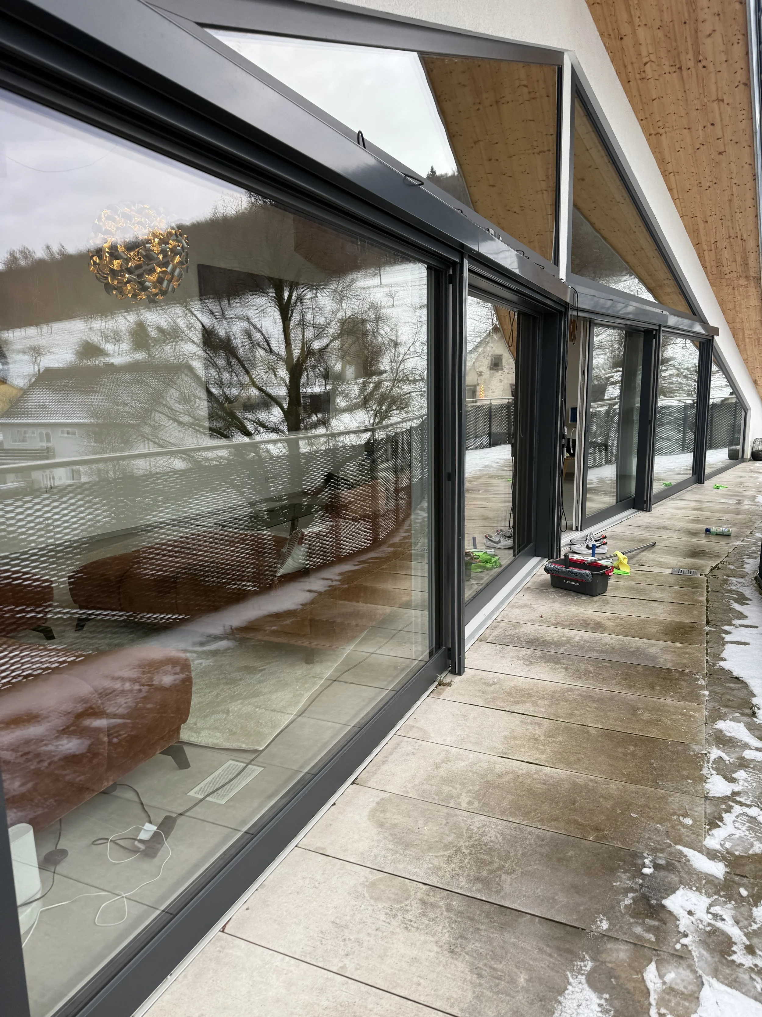 Modernes Haus mit großen Glasfenstern und Holzdecke, Blick auf einen Balkon im Winter mit Schneefall, Außenansicht eines Hauses mit Bäumen im Hintergrund, Werkzeuge auf dem Balkon.