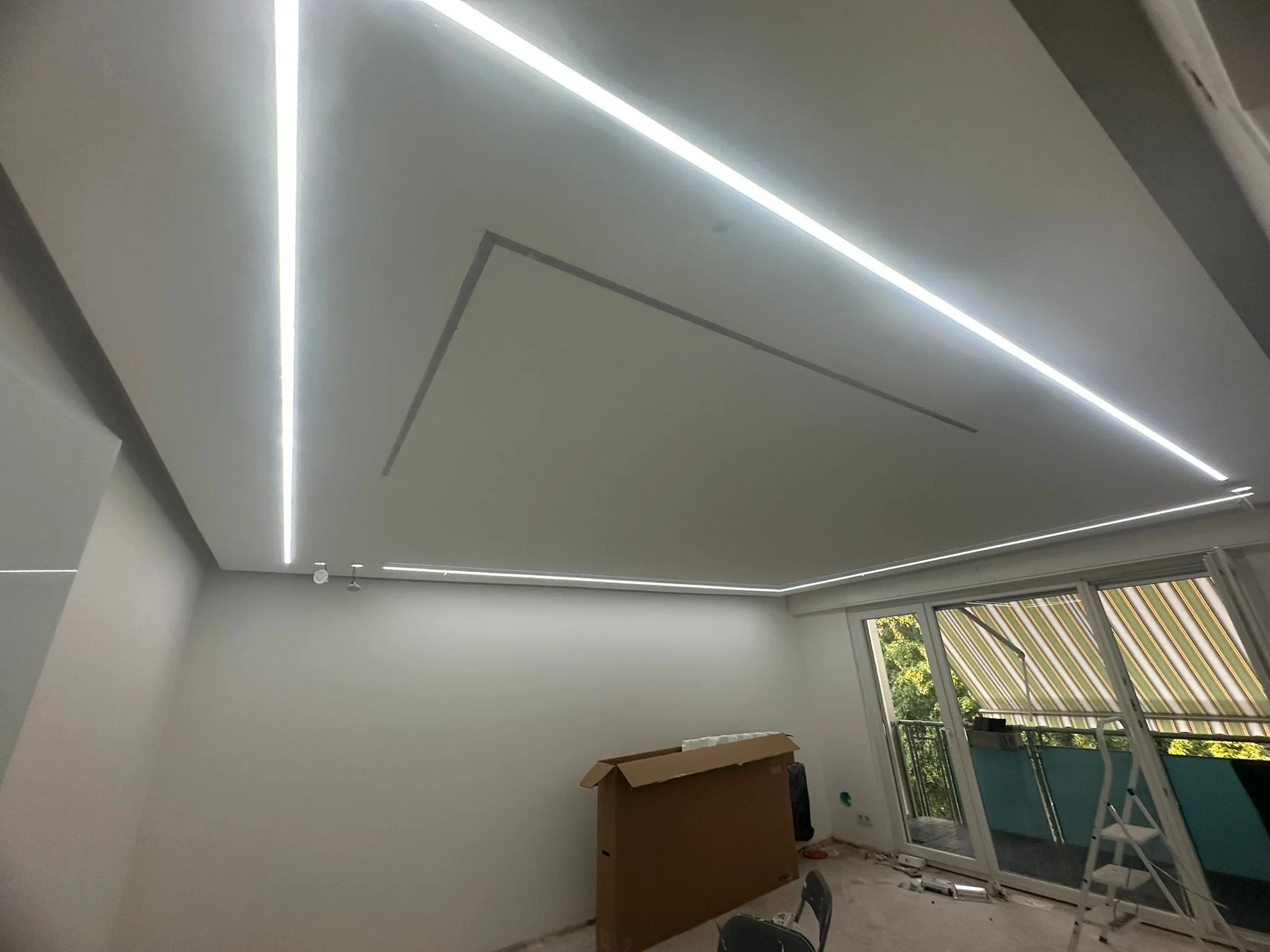 Moderner Raum mit weiß geklebter Decke, integrierten LED-Lichtstreifen, offene Fenster mit Balkon und Segeltuch-Markise im Hintergrund.
