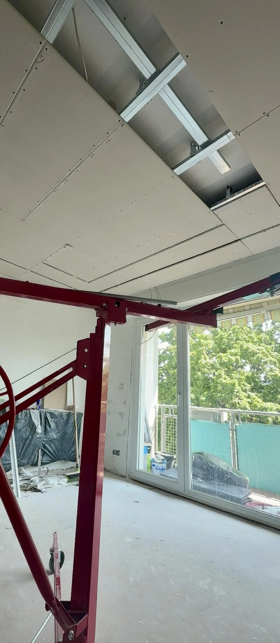 Innenaufnahme eines Zimmers während der Renovierung mit sichtbarer Decke, eine große Fensterfront und einen Balkon im Hintergrund. Im Zimmer steht ein rotes Baugerüst.