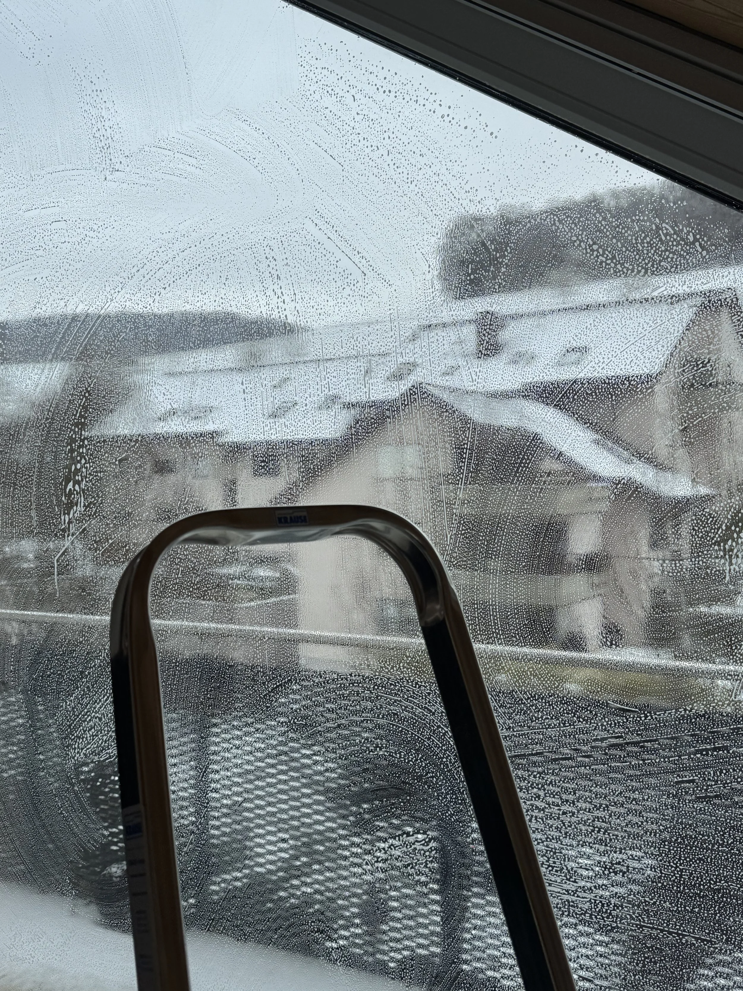 Fenster mit beschlagener Scheibe und Wassertropfen, Blick nach draußen auf ein silbrig glänzendes Landfahrzeug, auf dem sich eine helle Decke befindet.