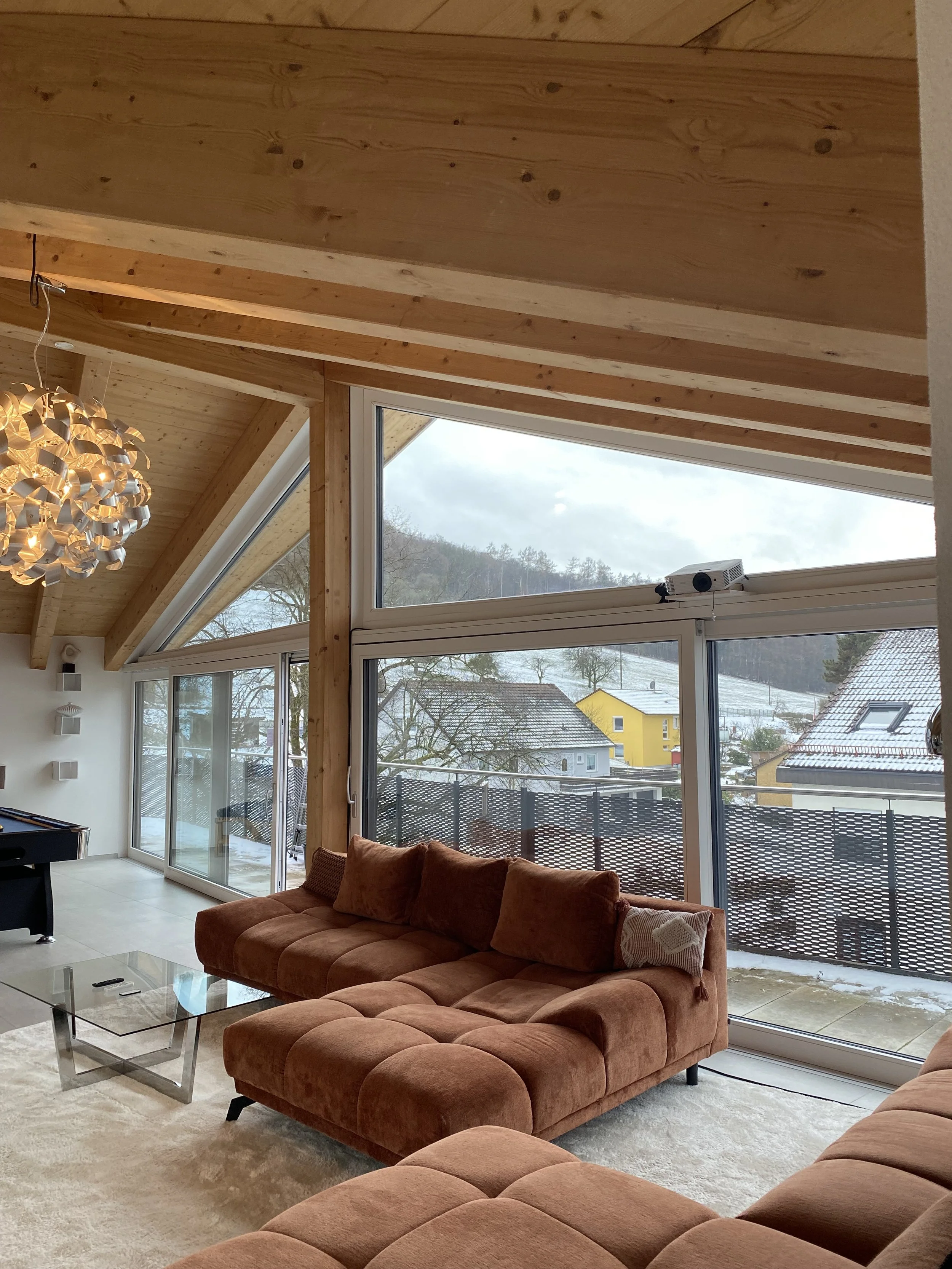 Wohnzimmer mit großen Fenstern, Couch, Glastisch, Holzdecke und Schneelandschaft im Hintergrund.