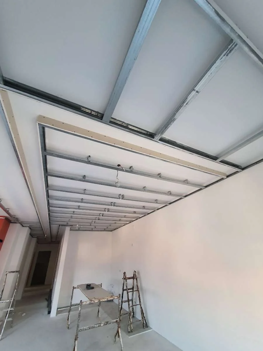 Innenraum eines Gebäudes im Bau mit Metallrahmen für eine abgehängte Decke, Leitern, Arbeiterwerkzeuge und leere Wände.