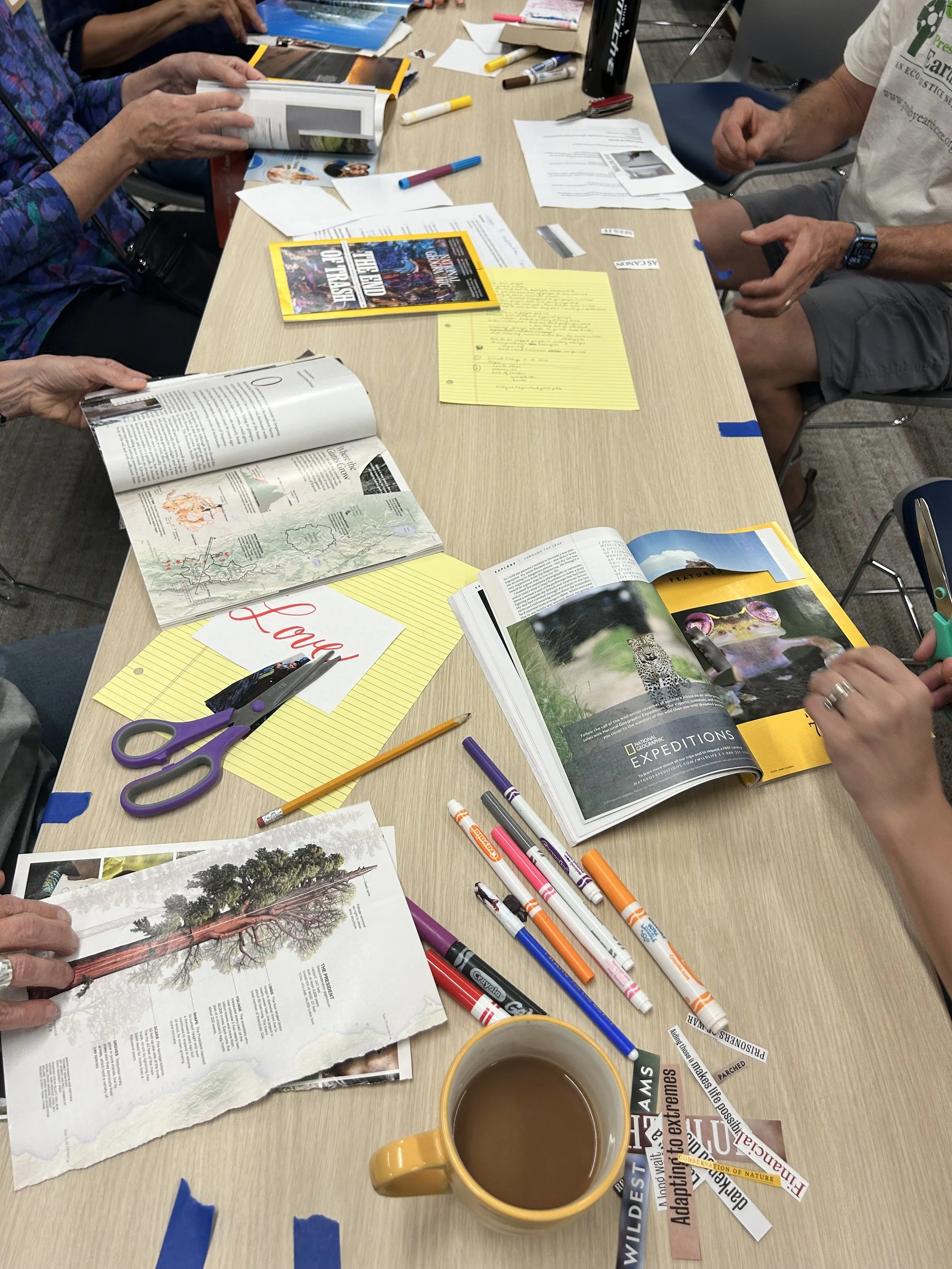 People seated around a table working on a communal craft, with magazines, pens, markers, scissors, paper, and a cup of coffee on the table. One paper is visible with calligraphy letters spelling "love."