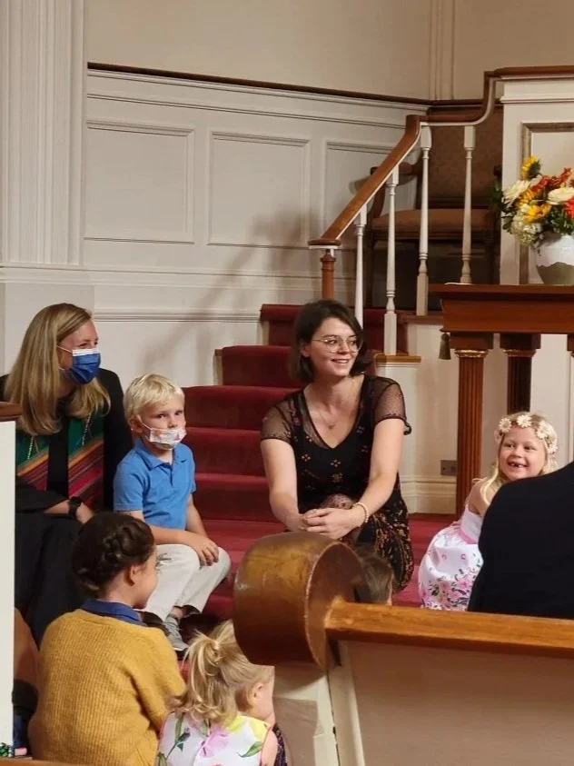 Group of children and adults sitting on church pews and stairs during a gathering or event, with a woman smiling and a young girl in a floral dress in the foreground.