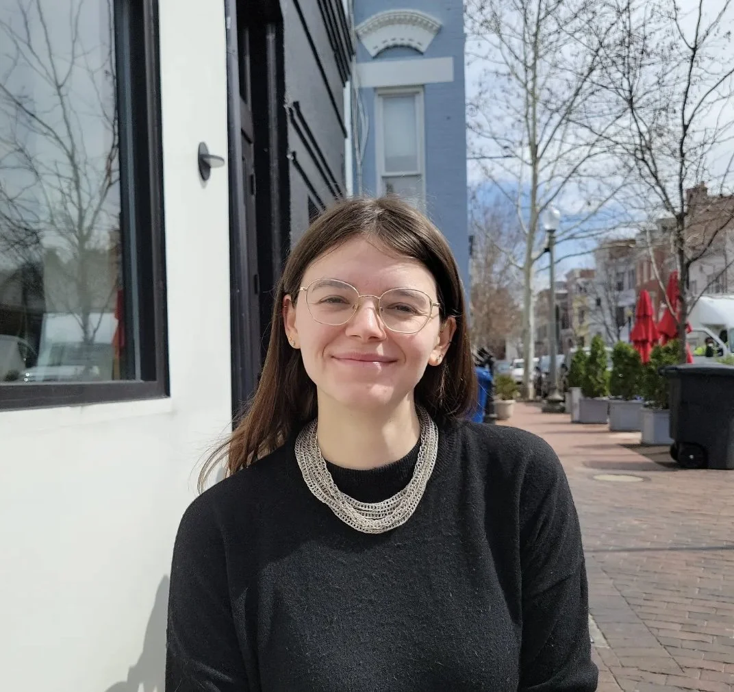 A young woman with glasses and a layered necklace smiling outdoors on a city street with buildings, trees, and red umbrellas in the background.