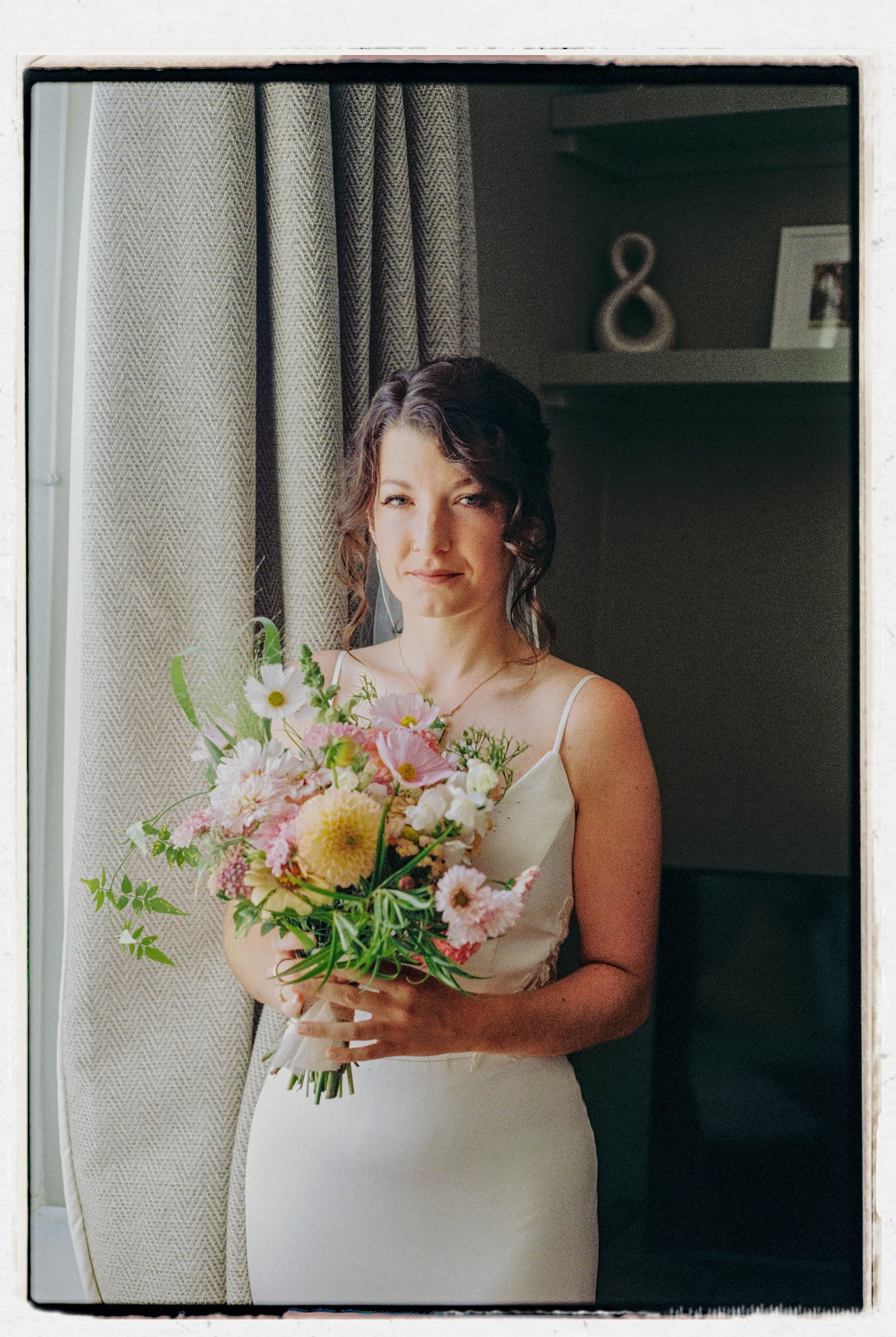 A woman in a wedding dress holding a bouquet of pink and white flowers, standing in front of curtains and a dark wall with a decorative item in the background.