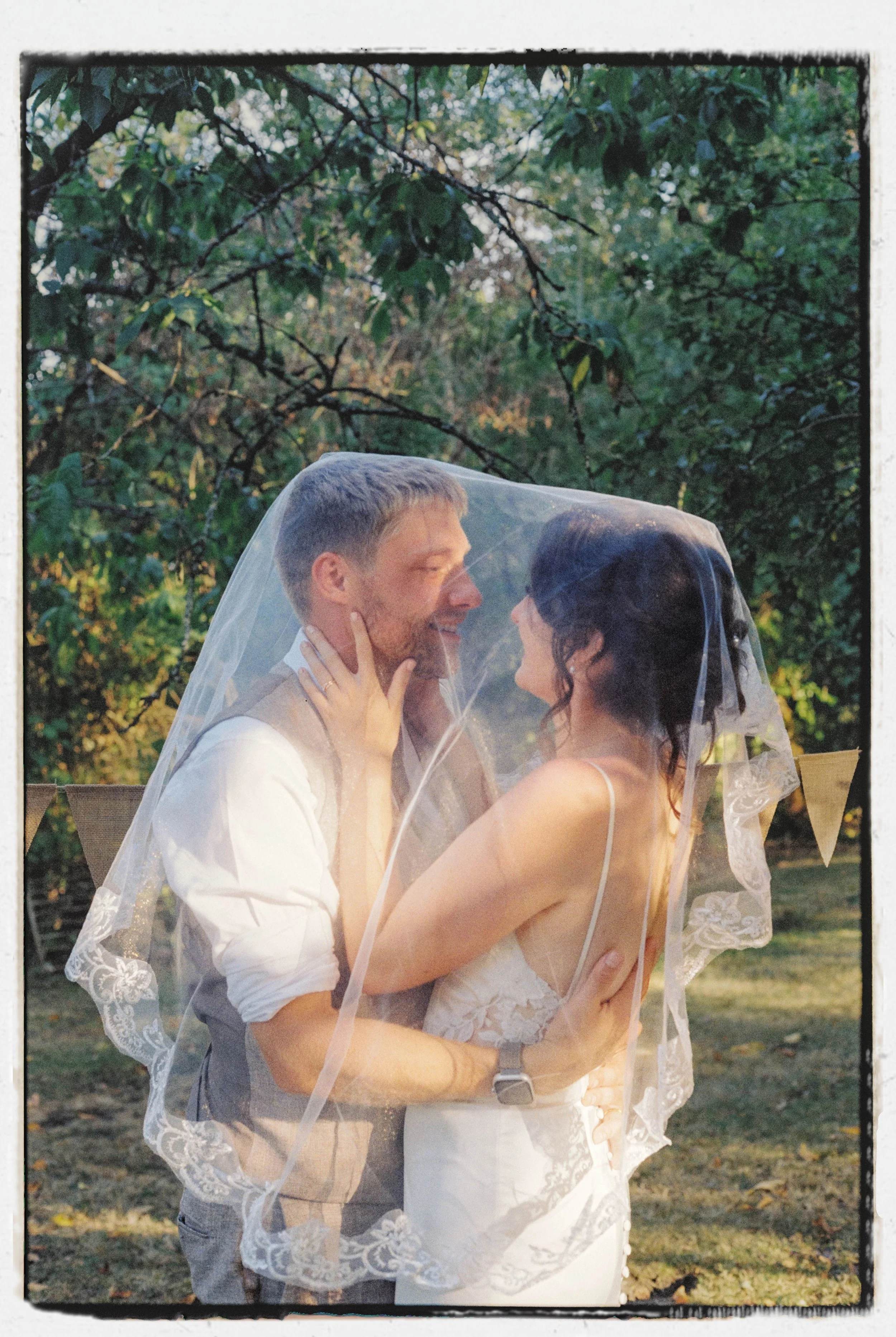 A newly married couple smiling at each other under a white veil outdoors, surrounded by trees with green leaves.