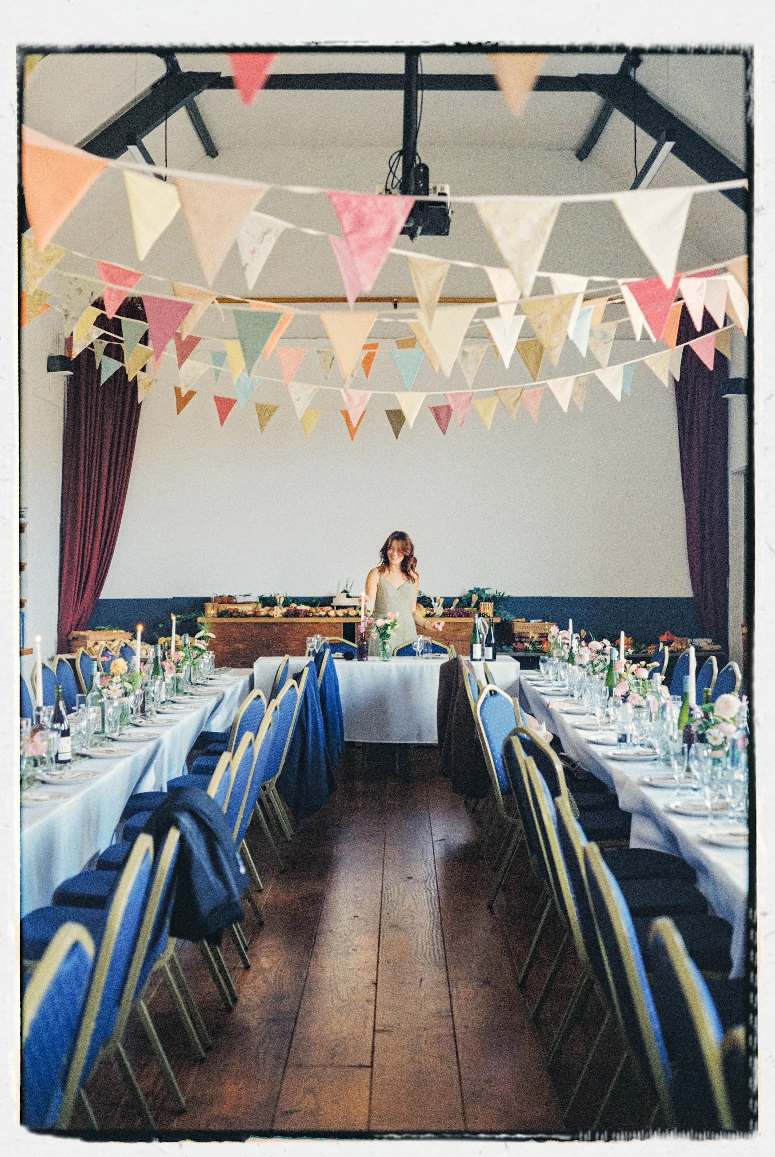 A decorated banquet hall with long tables set for a celebration, adorned with floral arrangements and candles, and colorful bunting hanging from the ceiling.