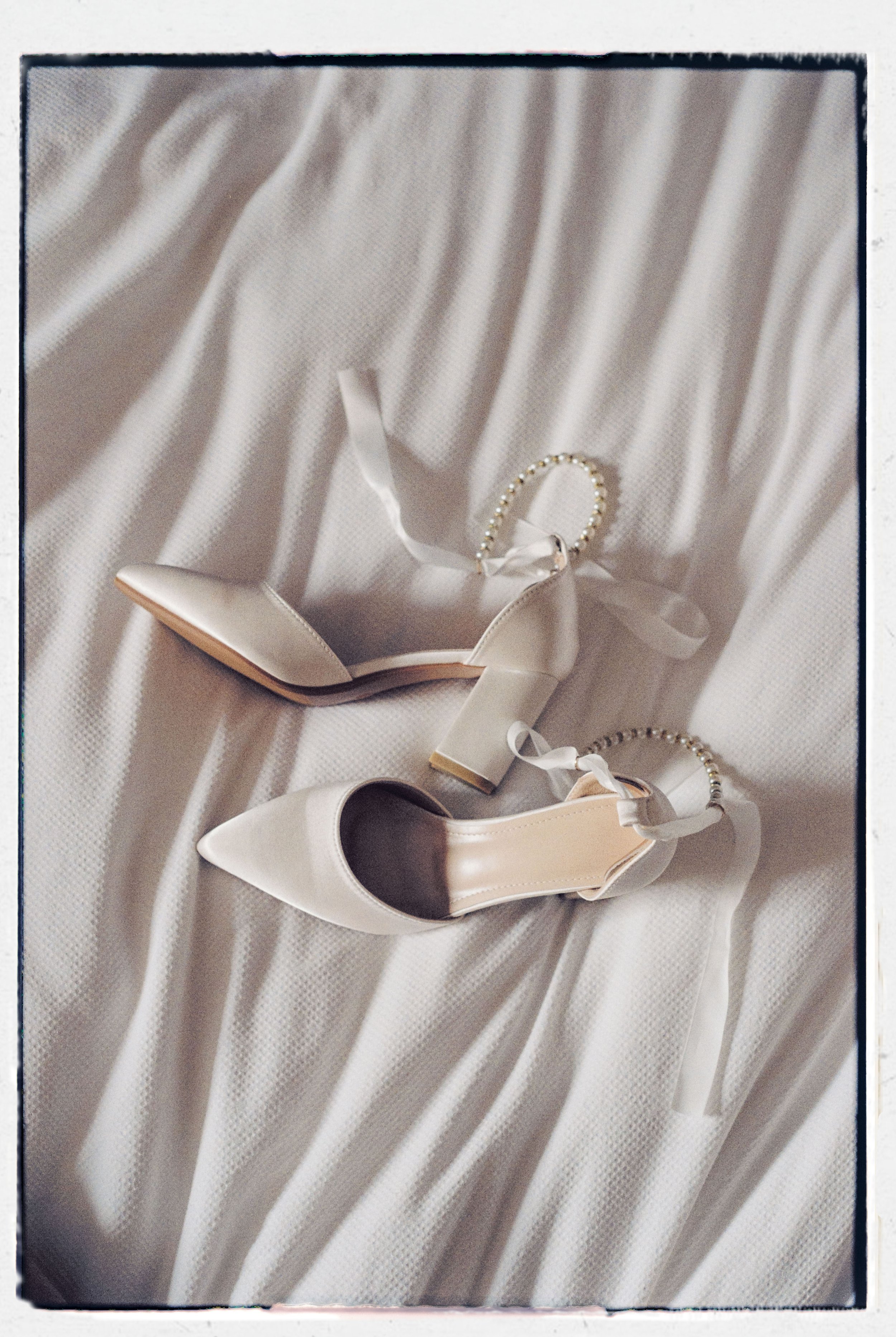 A pair of white high-heeled shoes with pointed toes and ankle straps, and a pearl bracelet, all placed on a white textured fabric background.