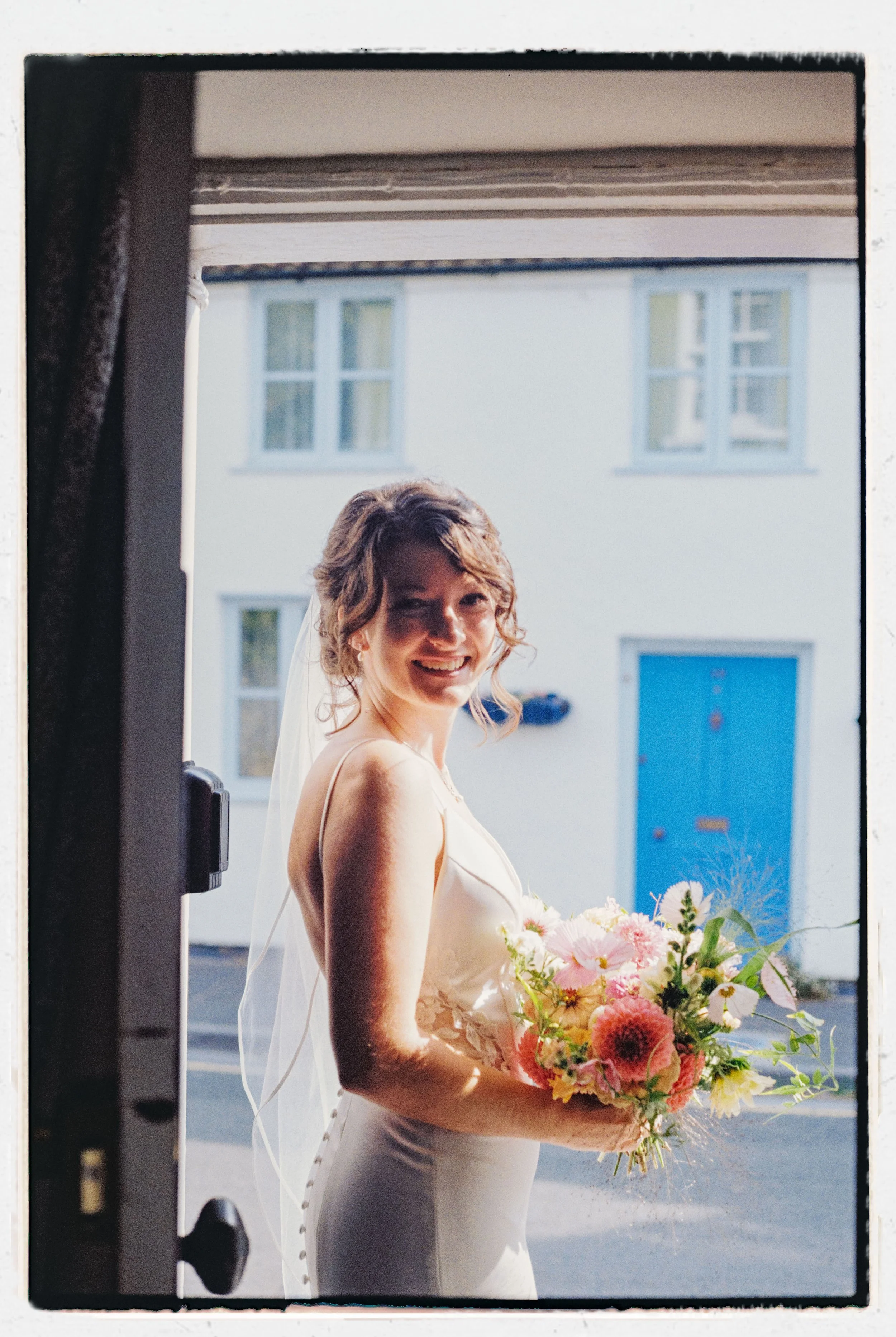 A bride standing outside holding a bouquet of pink and white flowers, smiling, near a building with blue windows and a blue door, with sunlight illuminating her face.