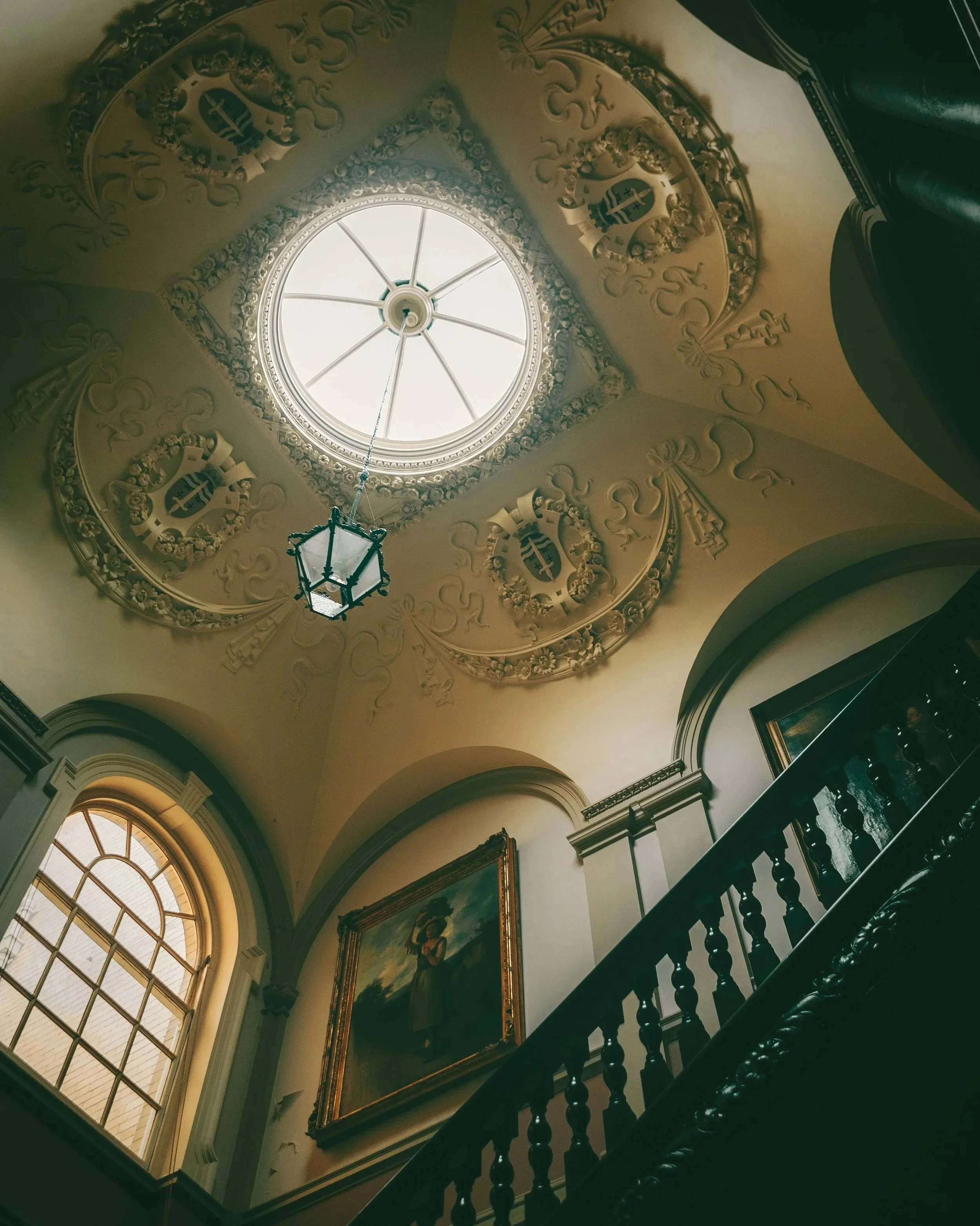 View of a grand ornate ceiling with a central skylight, decorative molding, a hanging lantern, large arched window, and framed painting on the wall near a staircase.