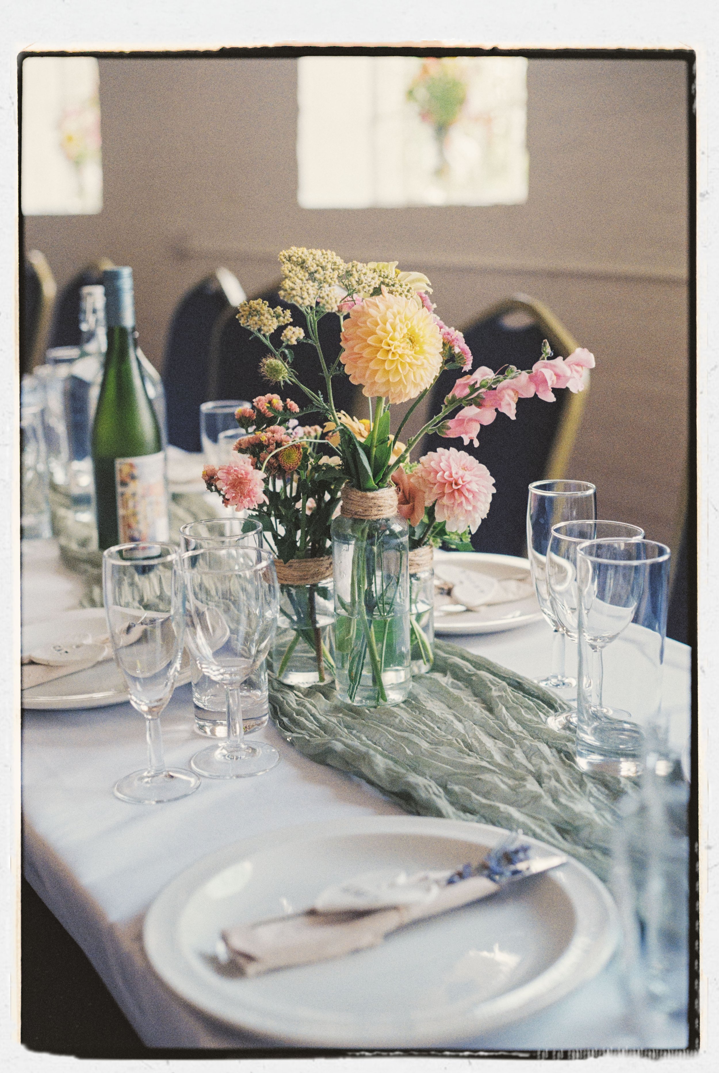 A dining table decorated with a bouquet of pastel-colored flowers in a glass jar, surrounded by wine bottles, glasses, plates, and napkins. The setting appears to be for a special occasion or dinner gathering.