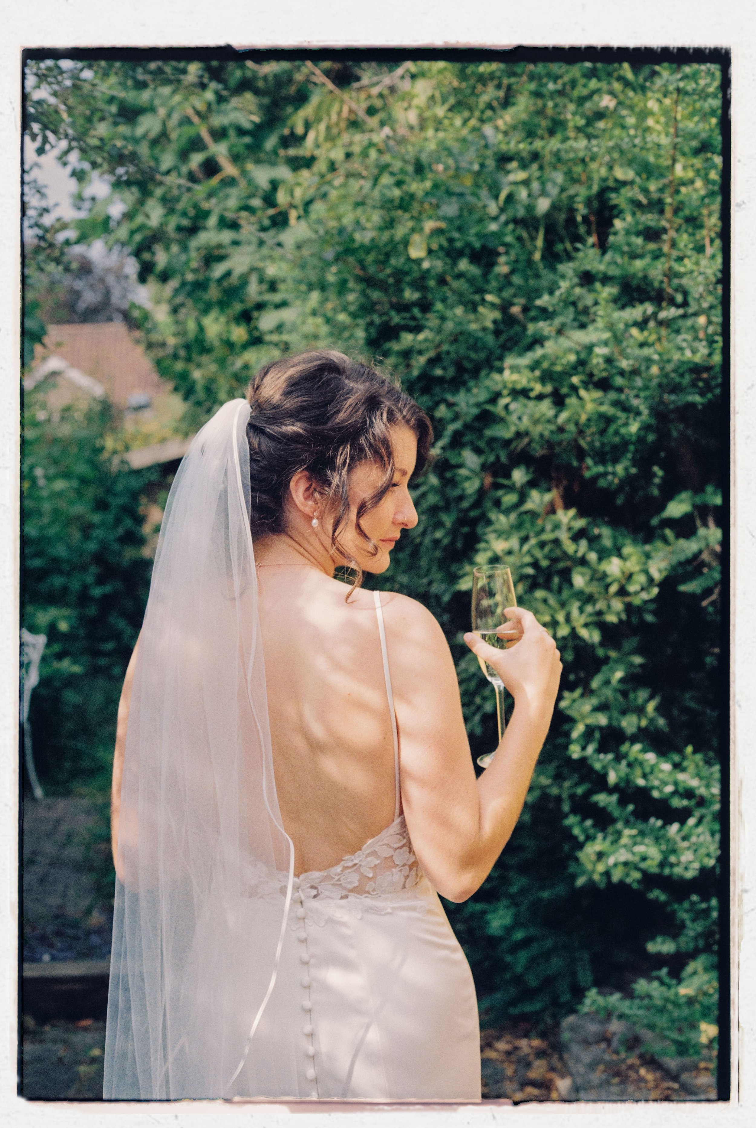 A bride with dark curly hair, wearing earrings and a sheer wedding dress with buttons on the back, holding a champagne flute, outdoors surrounded by green trees.
