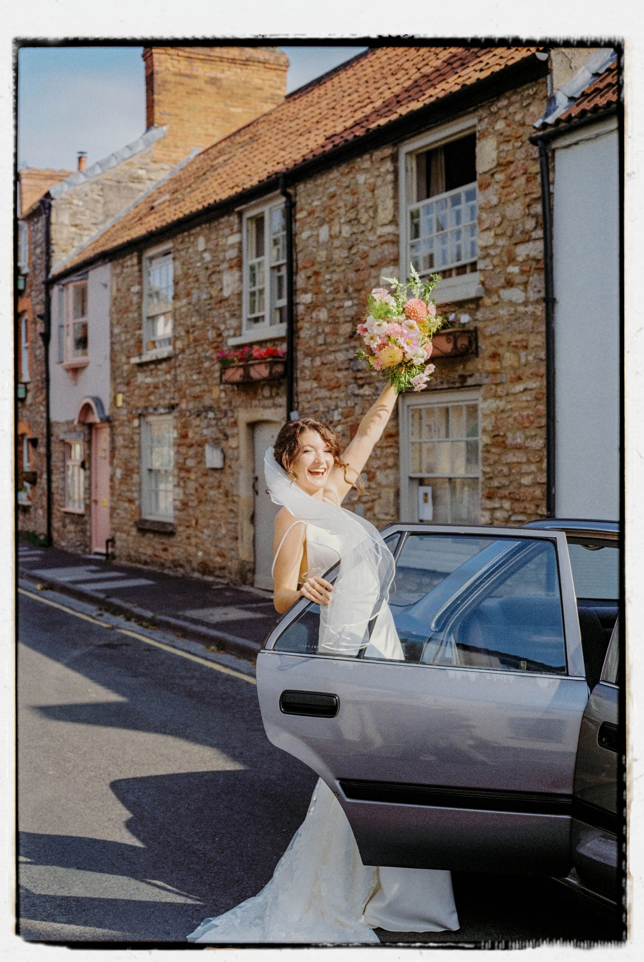 A joyful bride in a white wedding dress with a veil, holding a bouquet of pink and white flowers, standing next to a car with its door open on a street lined with brick houses.