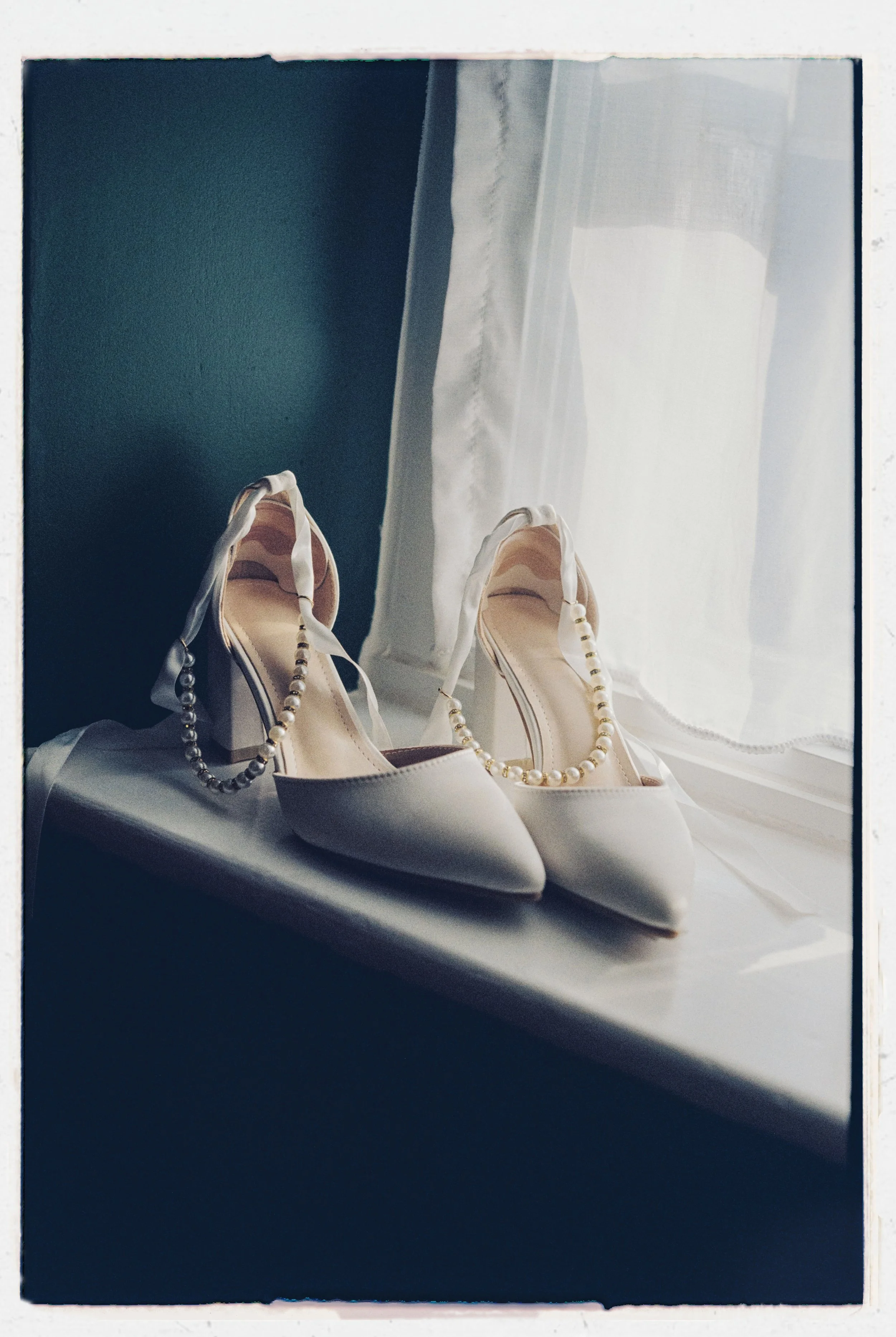 A pair of white high-heeled shoes with pearl necklaces draped over them, placed on a white surface near a window with sheer white curtains.