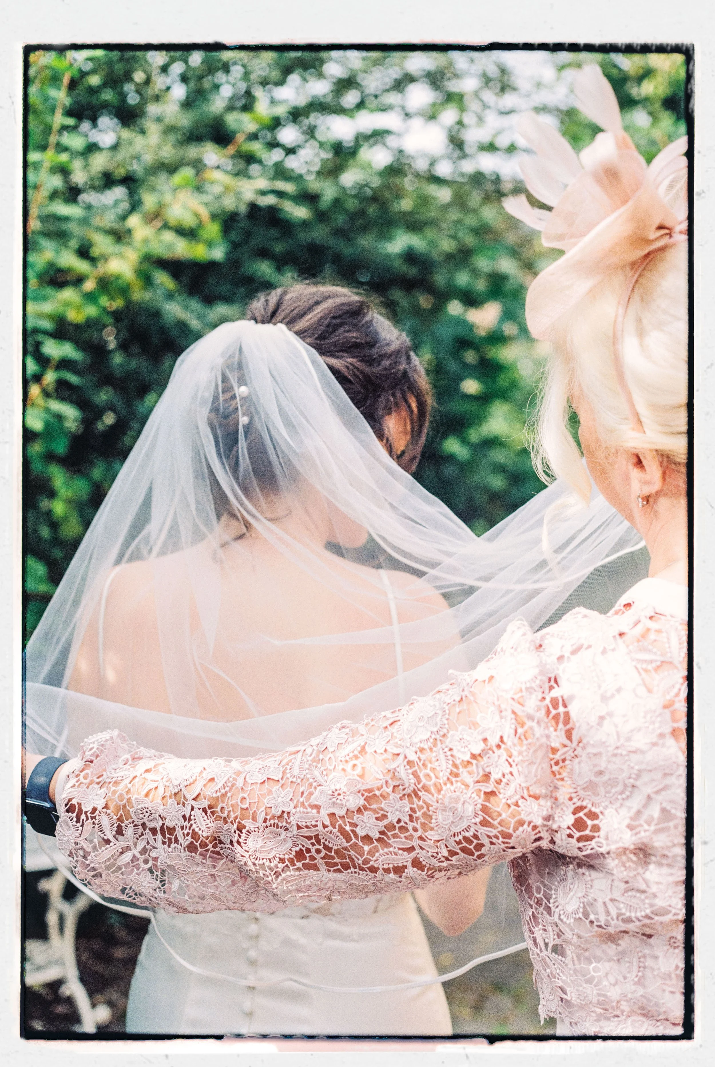 Close-up of a bride wearing a white dress and veil with another woman in a pink lace dress, outdoors with green trees in the background.