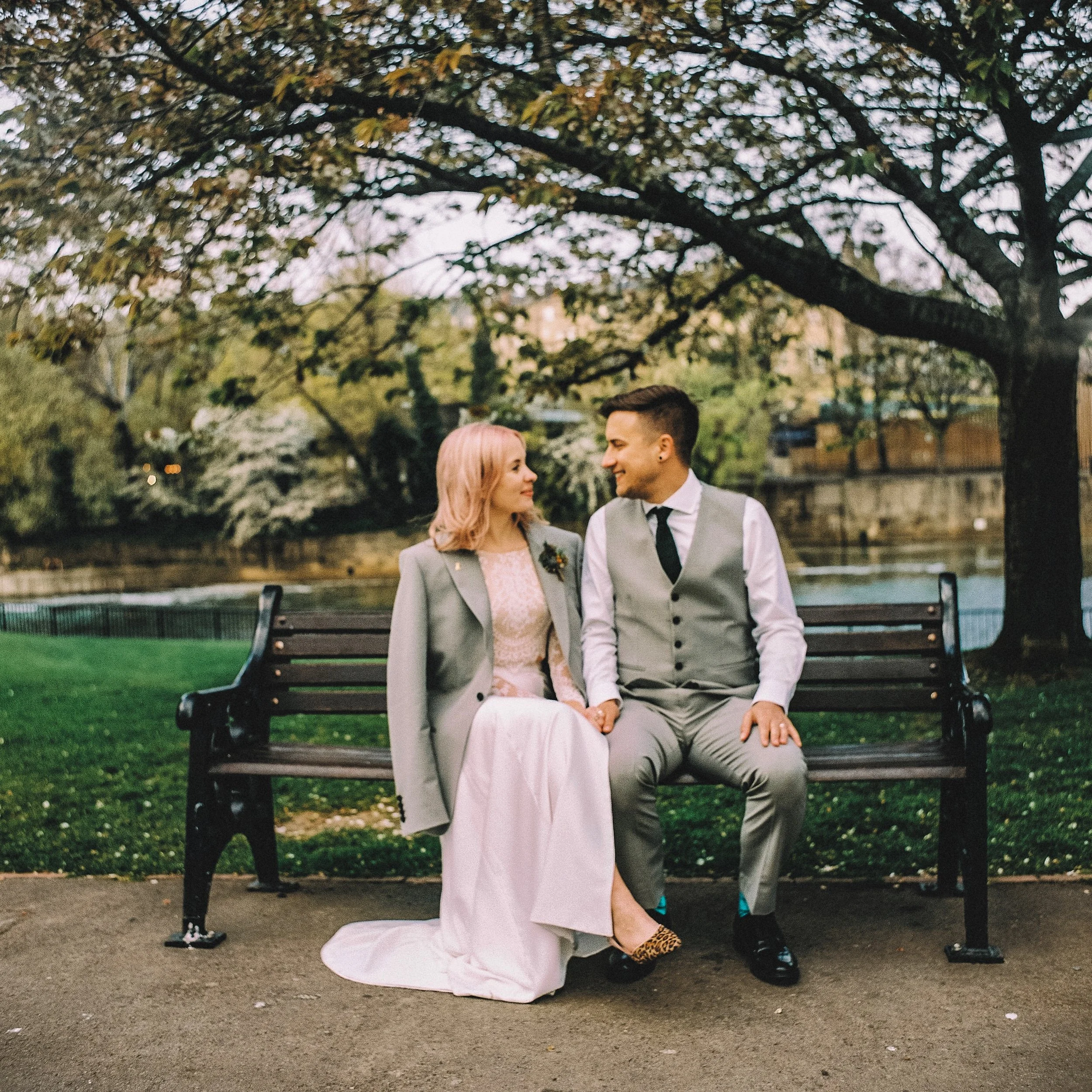 A couple sits on a park bench under a large tree, looking at each other and smiling. The woman is wearing a pink dress and gray blazer, and the man is in a gray vest and pants with a white shirt and black tie. The scene is set outdoors near water with greenery in the background, possibly during spring.