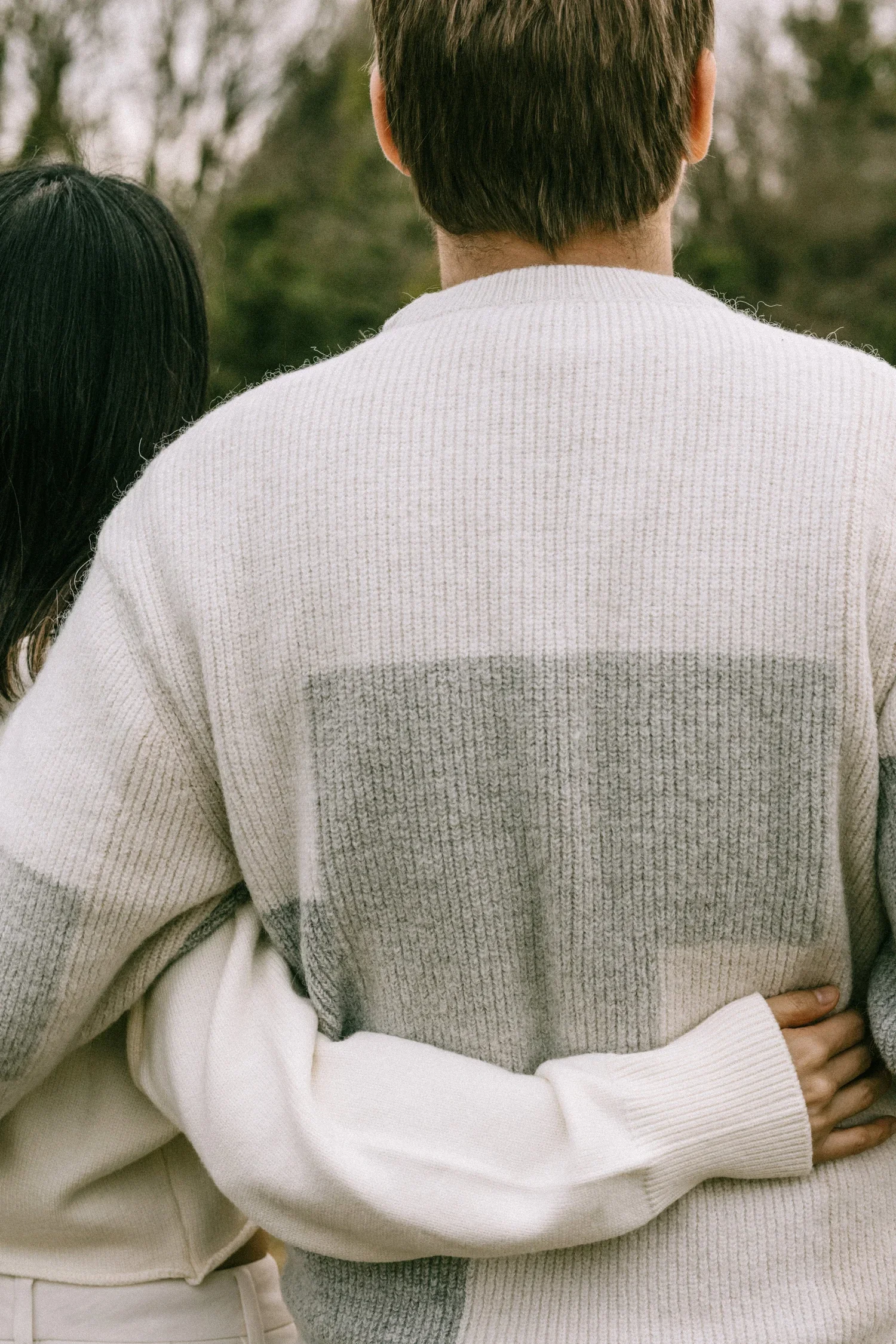 A person in a cream and gray colorblock sweater with another person's arm around their waist, outdoors with blurred trees in the background.