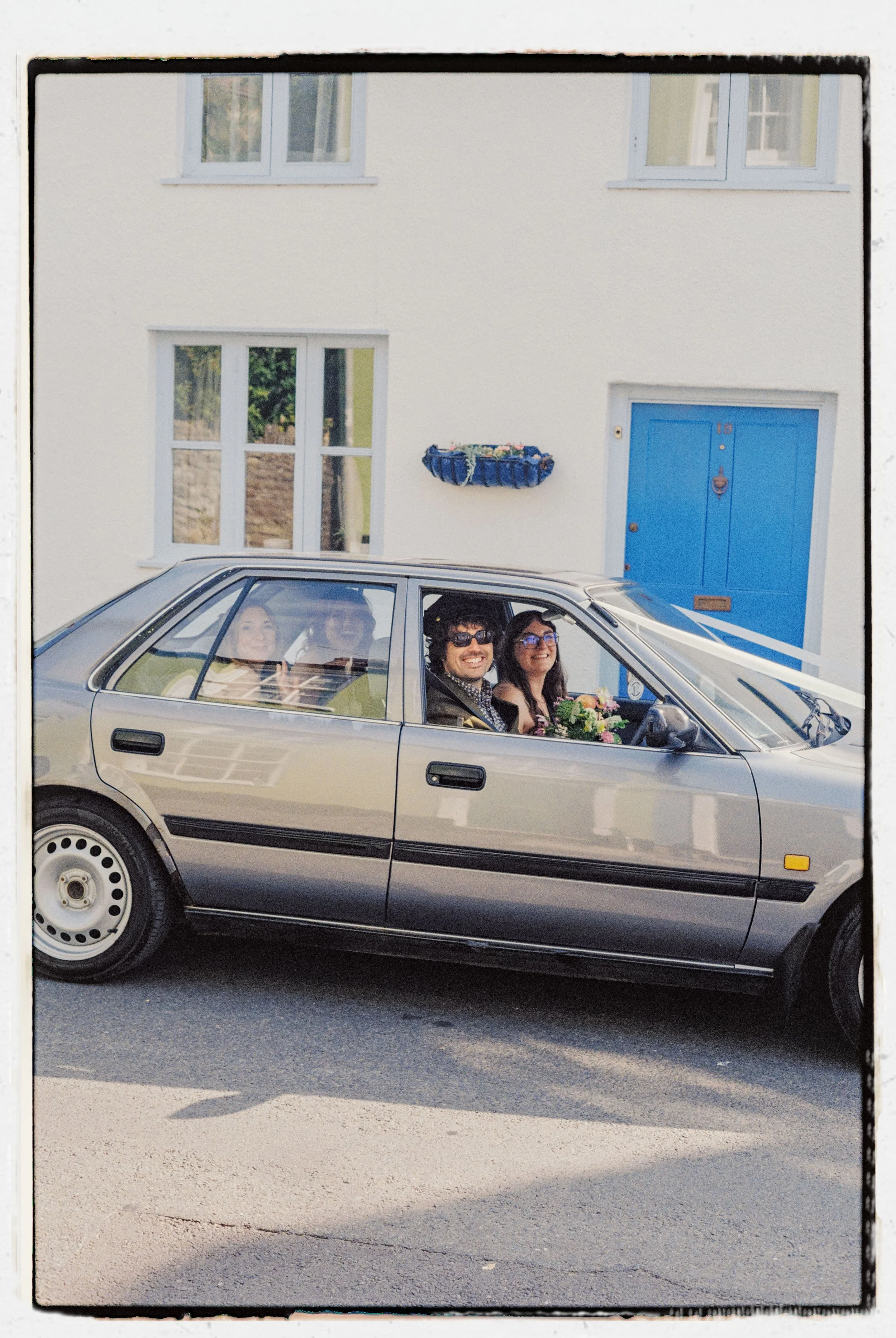 Four people sitting inside a silver car, visible through the windows, parked in front of a white house with a blue door and window boxes