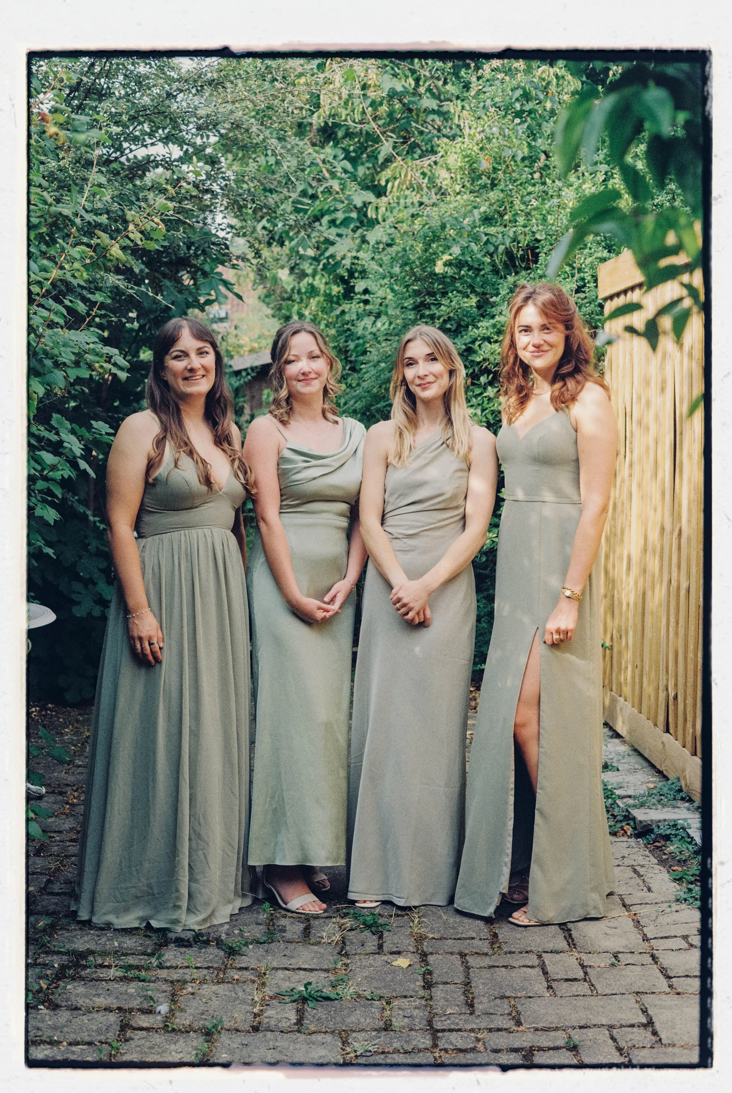 Four women standing outdoors on cobblestone pavement, surrounded by greenery and a wooden fence, wearing matching sage green dresses and smiling.