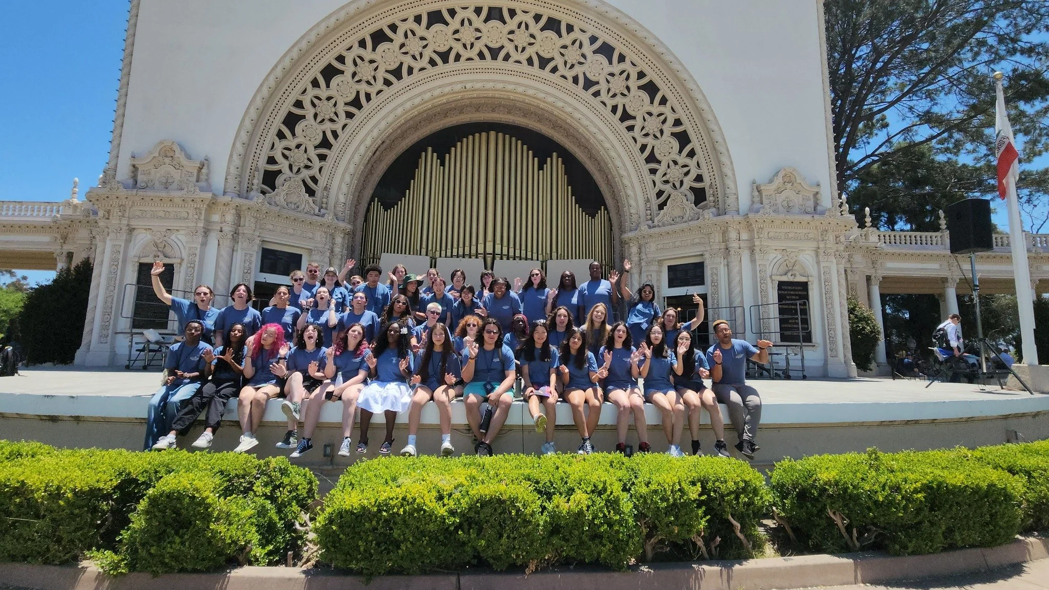 A large group of people wearing matching blue shirts sitting and standing in front of a building with ornate architectural details, pipes, and an arch with decorative patterns, under a clear blue sky.