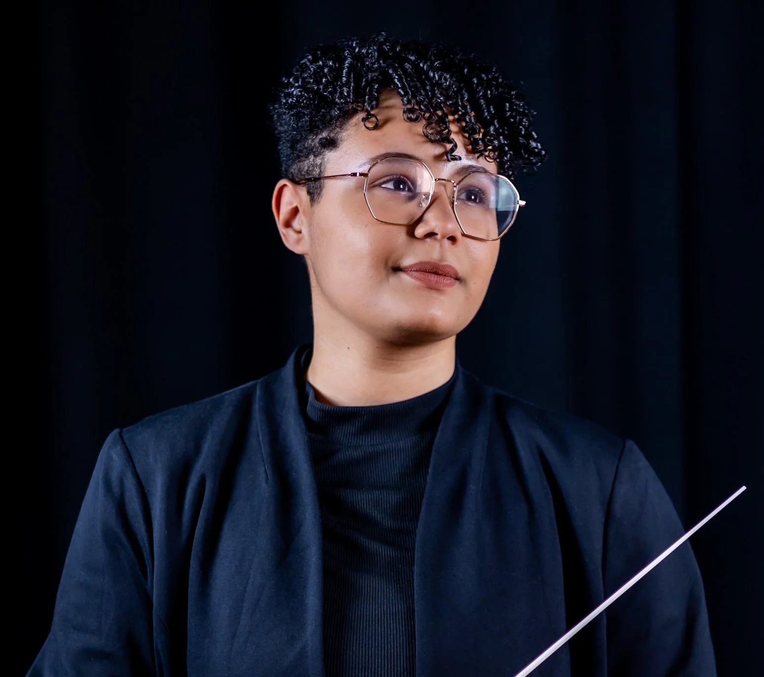 A person with short, dark curly hair and glasses, wearing a black jacket and turtleneck, against a black background.