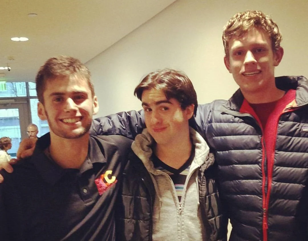 Three young men standing together indoors, smiling at the camera; two wearing dark puffer jackets and the one in the middle wearing a black and white hoodie.