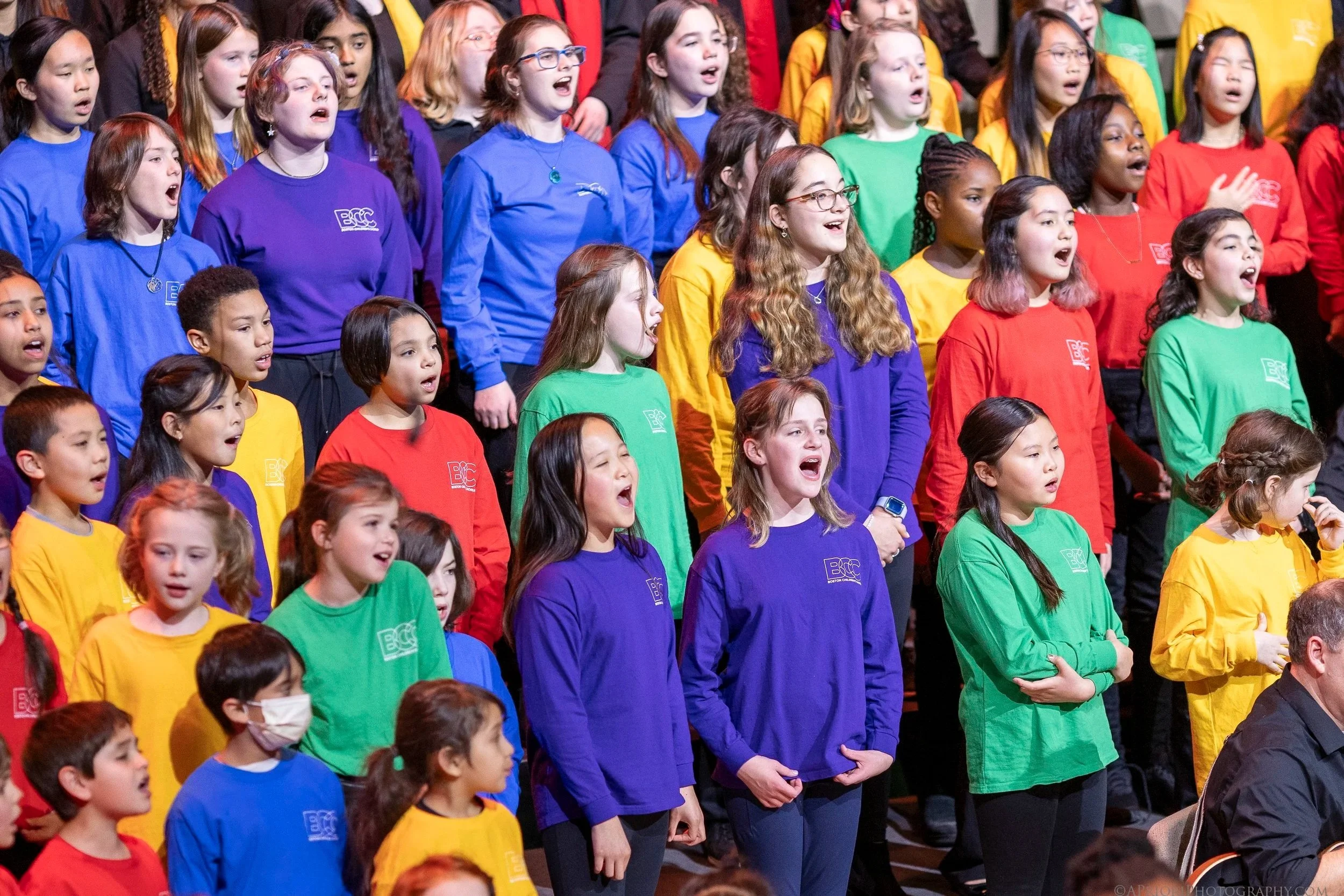 A large choir of children singing, wearing colorful shirts in blue, purple, green, yellow, and red, with some children wearing glasses or face masks, during a performance or rehearsal.