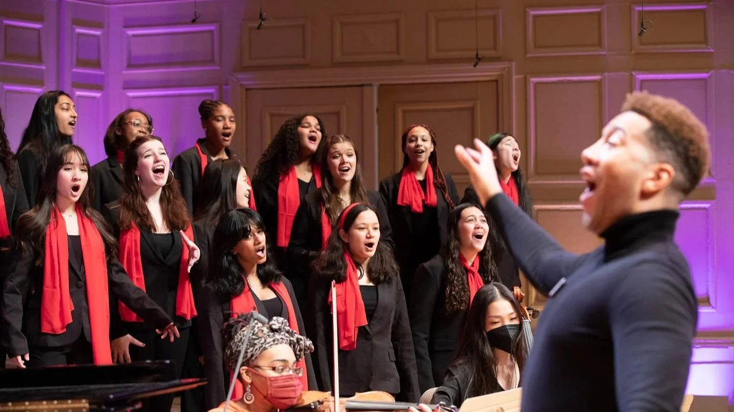 A choir of young women singing under the direction of a conductor in a concert hall with purple lighting.