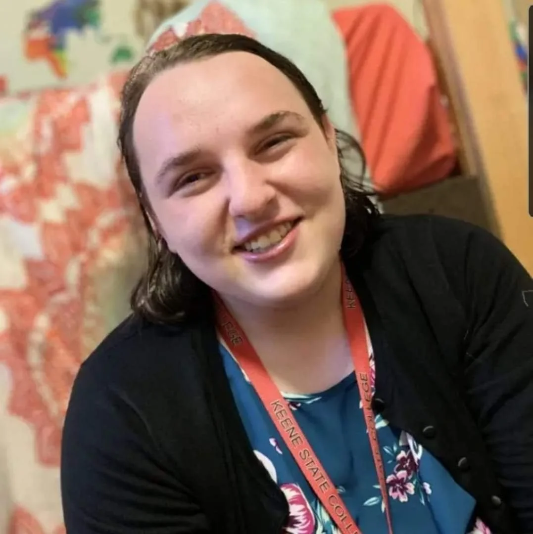 A young person with shoulder-length dark hair smiling at the camera, wearing a black jacket and a blue floral shirt, with a red lanyard around their neck that says 'Keene State College', and a colorful quilt or tapestry in the background.