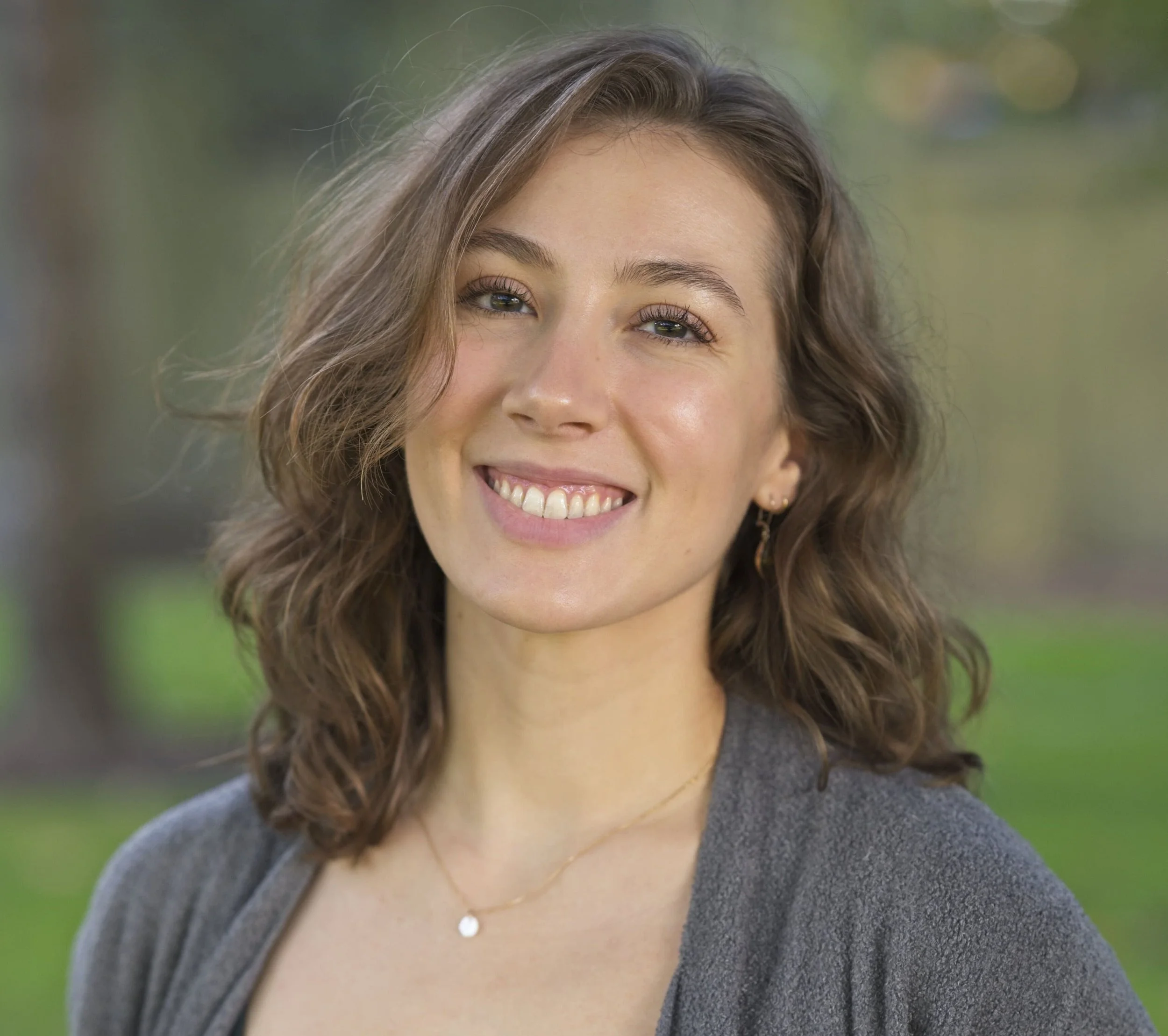 Close-up of a woman with long wavy brown hair smiling outdoors on a cloudy day, wearing a necklace with a small pendant and a dark gray sweater, with blurred green and yellow background.