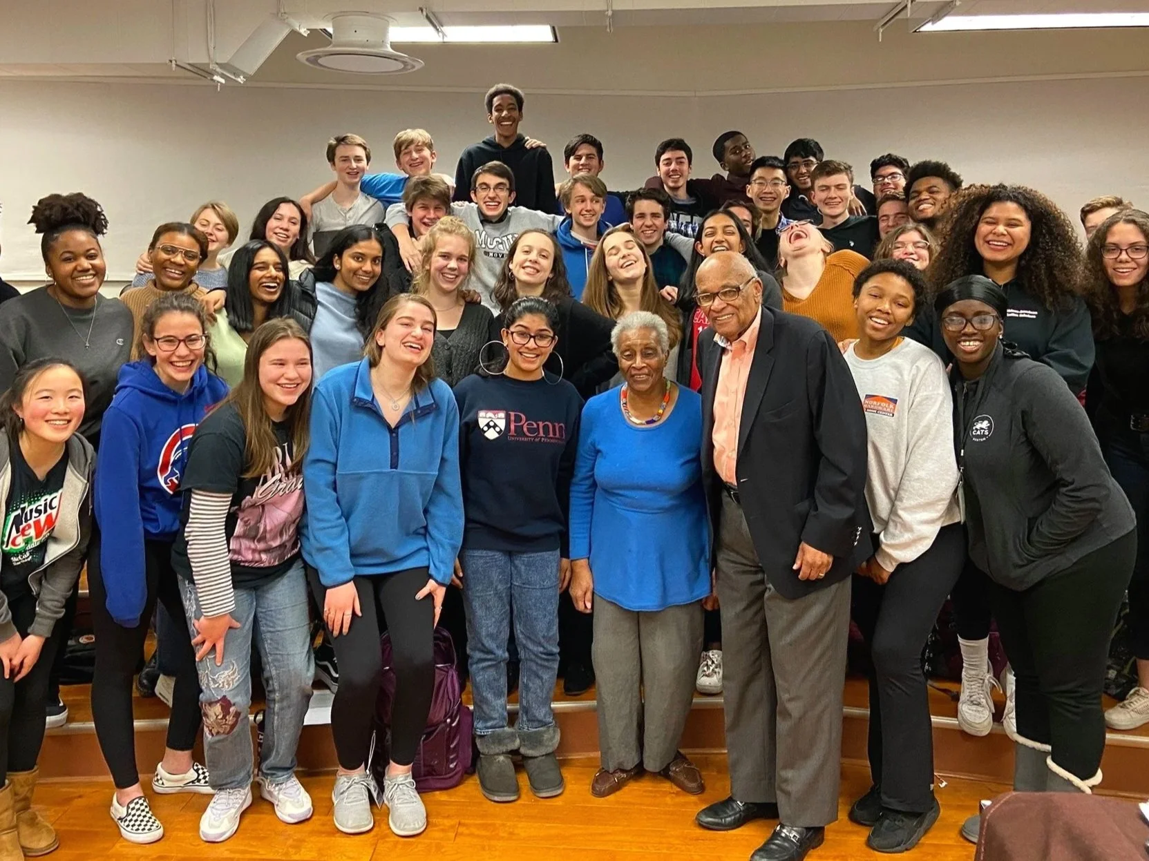 A large group of young people and three older adults gathered indoors for a group photo, smiling and laughing. The background is a plain wall and the group is diverse in ethnicity and dressed casually.