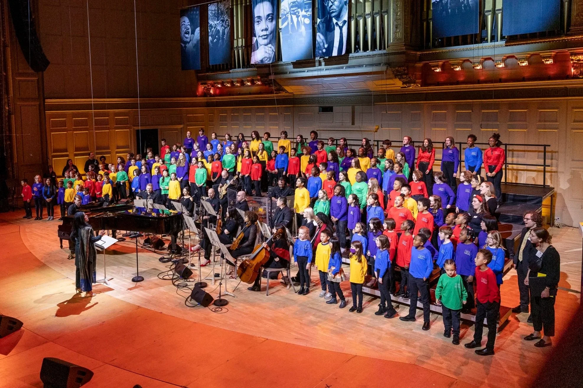 Children in colorful shirts standing on stage in a concert hall, performing with an orchestra, with large black and white portraits projected above them.