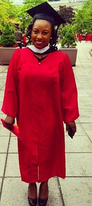 A woman in a red graduation gown and mortarboard cap standing outdoors during graduation ceremony.