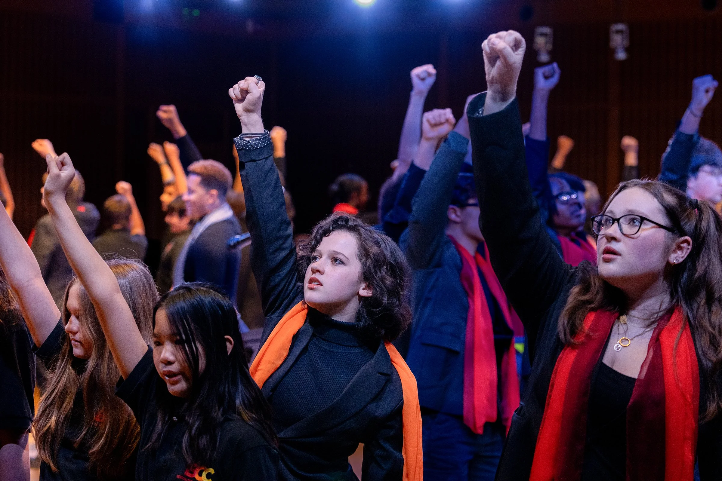 A crowded indoor event with many young people raising fists in the air, some with backpacks and various hairstyles, under dim lighting.