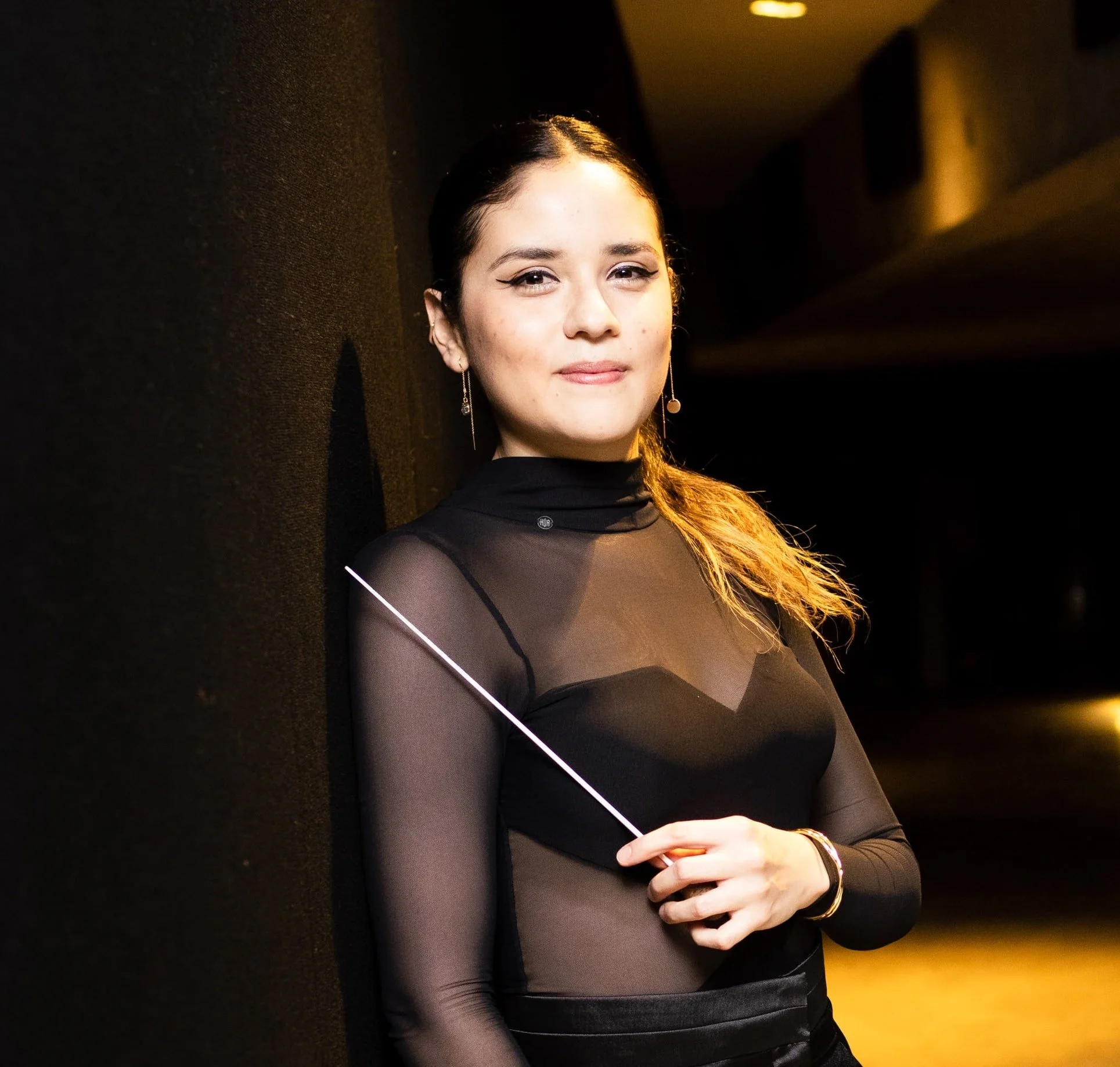 A woman with dark hair pulled back, wearing a black sheer top with long sleeves and earrings, standing against a dark background with yellowish lighting.