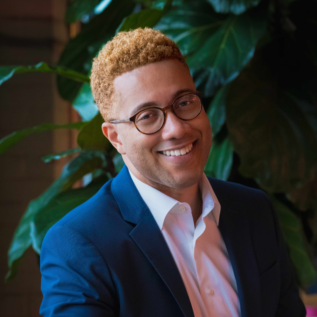 A smiling man with light brown curly hair, glasses, wearing a white shirt and dark suit jacket, standing in front of large green plants.