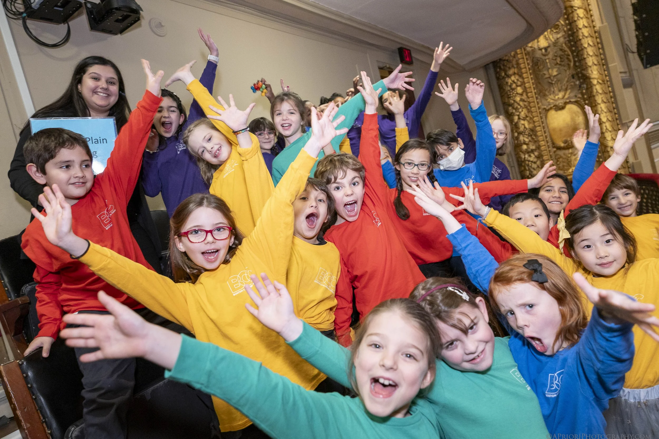 A group of children of diverse ethnicities wearing colorful shirts, smiling, and raising their hands in a celebration or event setting.