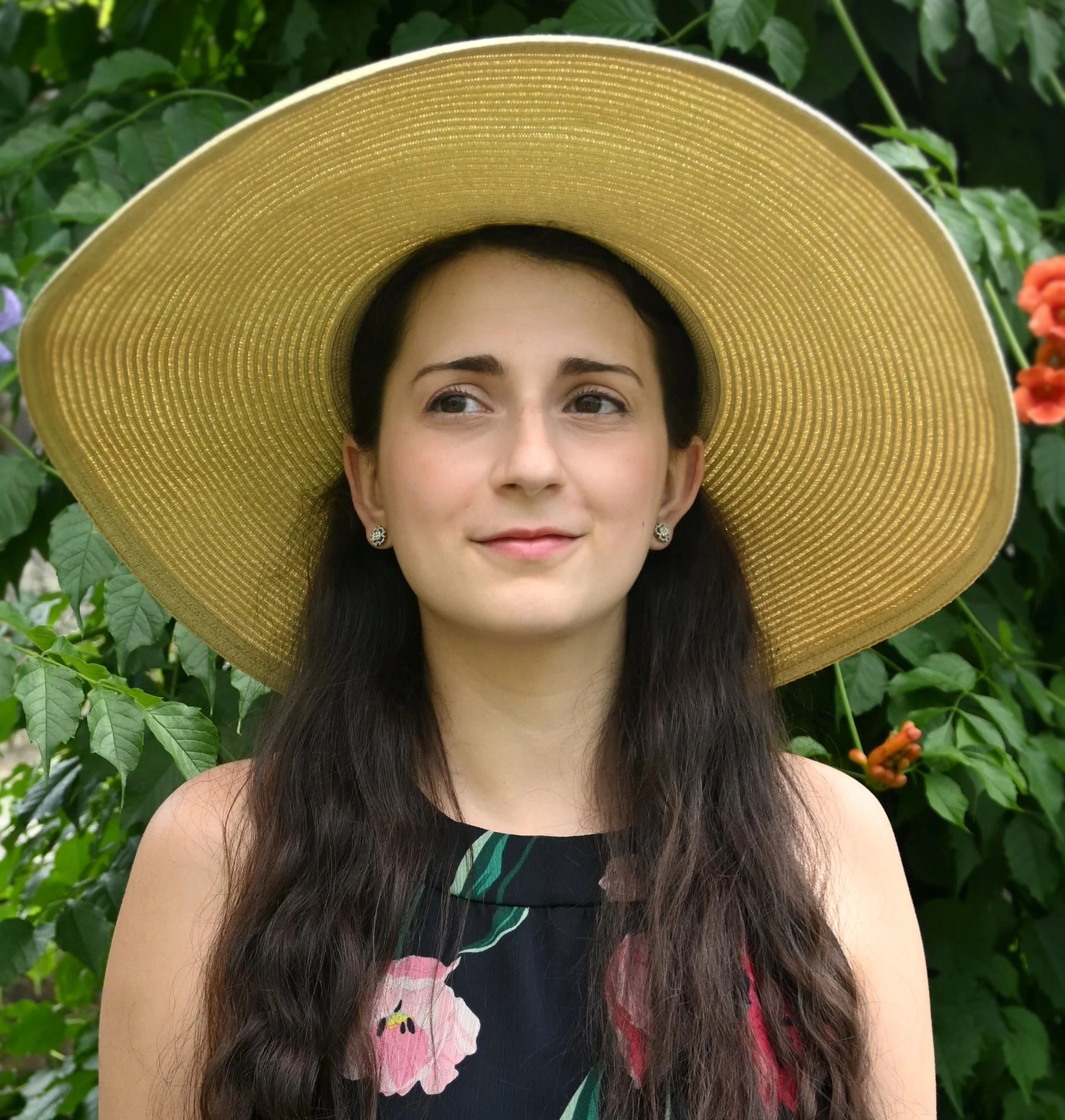 A woman wearing a wide-brimmed straw hat and a sleeveless dress with floral patterns stands outdoors among green leaves and colorful flowers.
