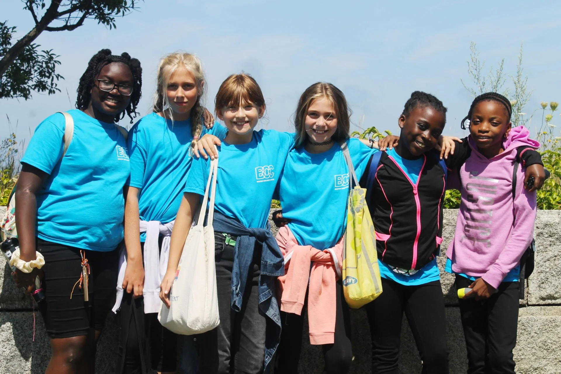 Group of seven young girls standing outdoors on a sunny day, smiling, with their arms around each other. They are wearing matching blue T-shirts, with some carrying backpacks and tote bags, dressed casually for an outdoor activity.