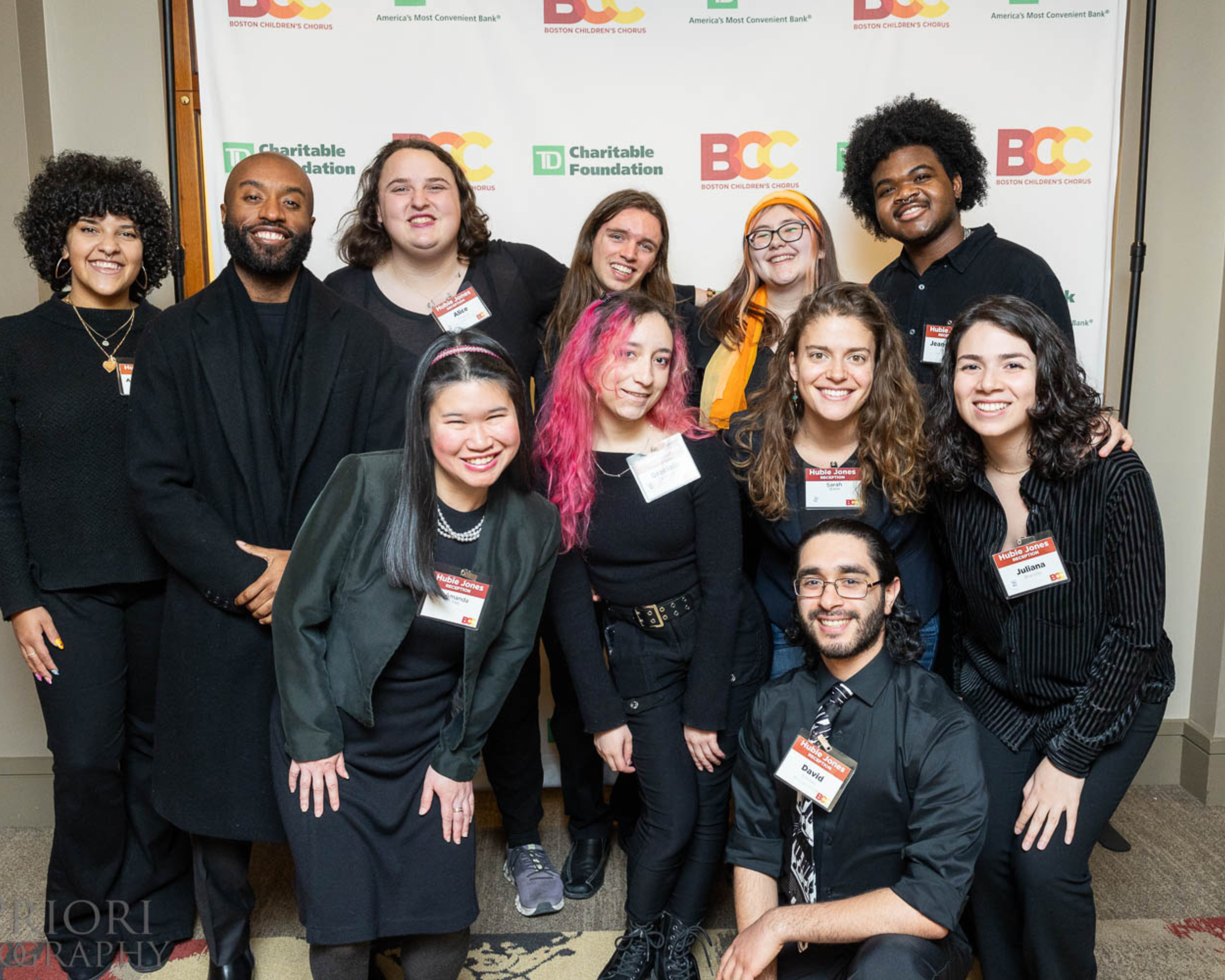 Group of diverse young adults smiling at an indoor event, with a banner displaying logos of Boston Children's Chorus, TD Charitable Foundation, and other sponsors behind them.