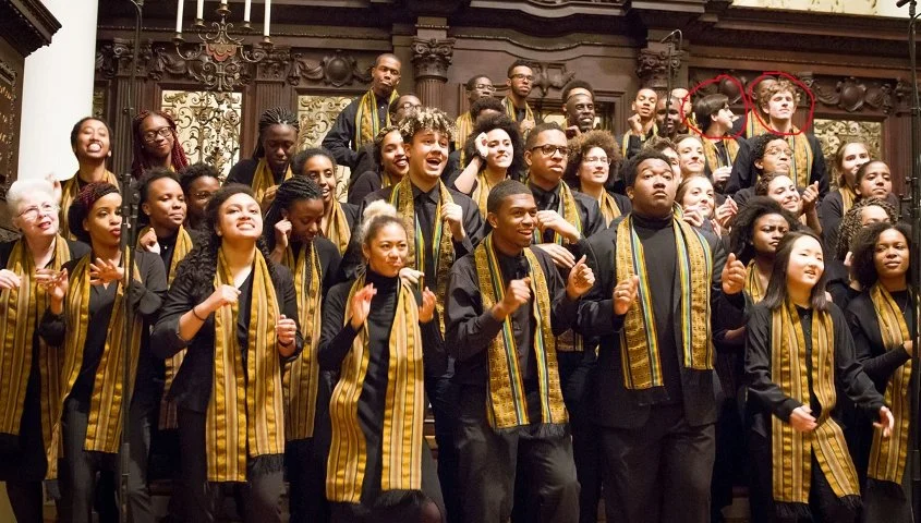 A diverse choir of young men and women in black outfits with yellow and black striped scarves, standing on a stage with ornate woodwork in the background, performing or celebrating.