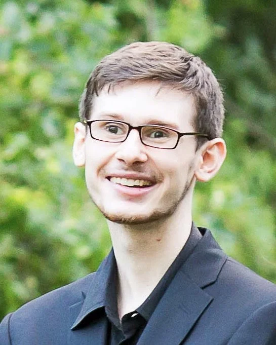 A young man with glasses and short brown hair, smiling, wearing a dark blazer, outdoors with trees in the background.