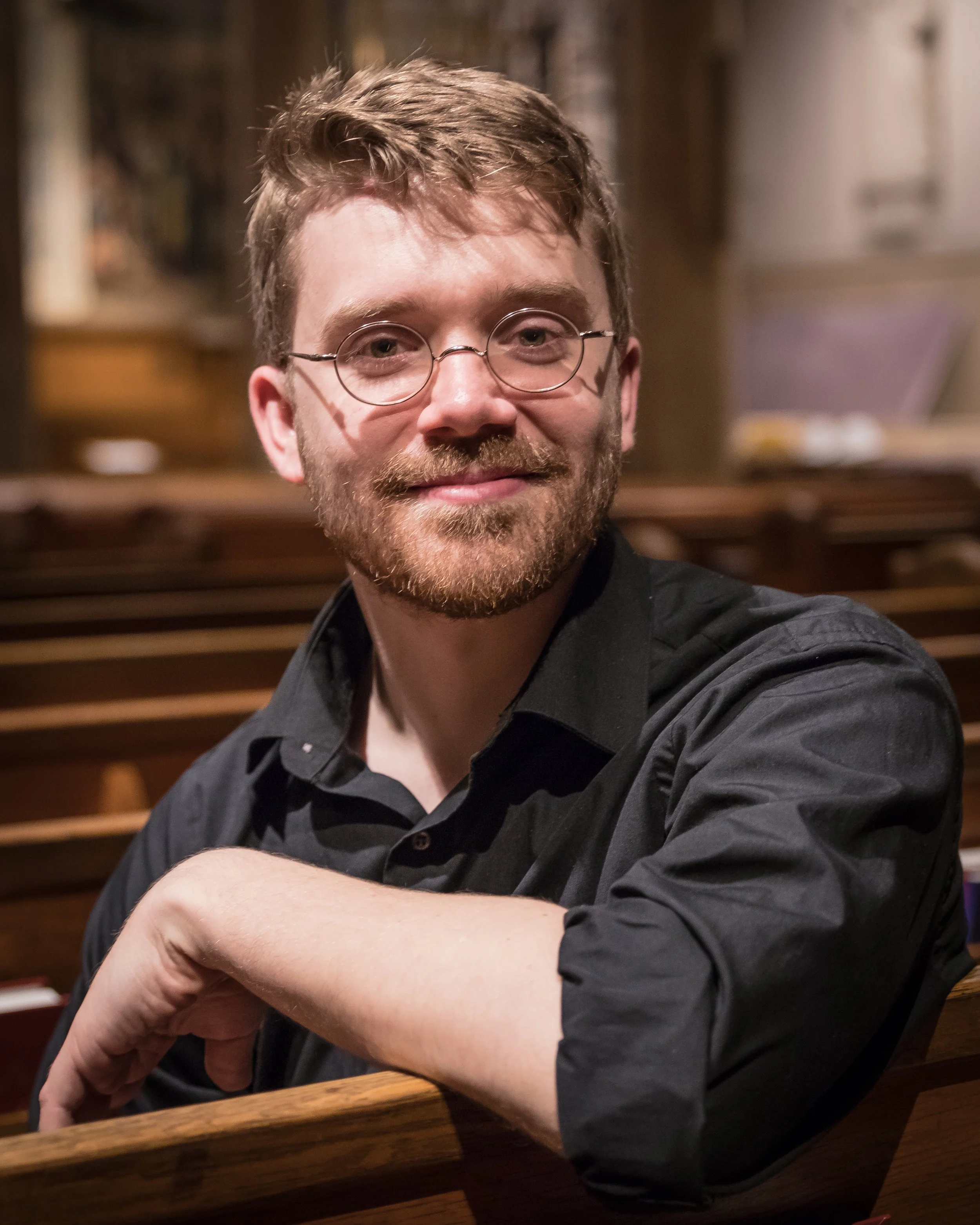 A man with glasses, brown hair, and a beard sitting at a wooden table, smiling in a warmly lit room with wooden walls.