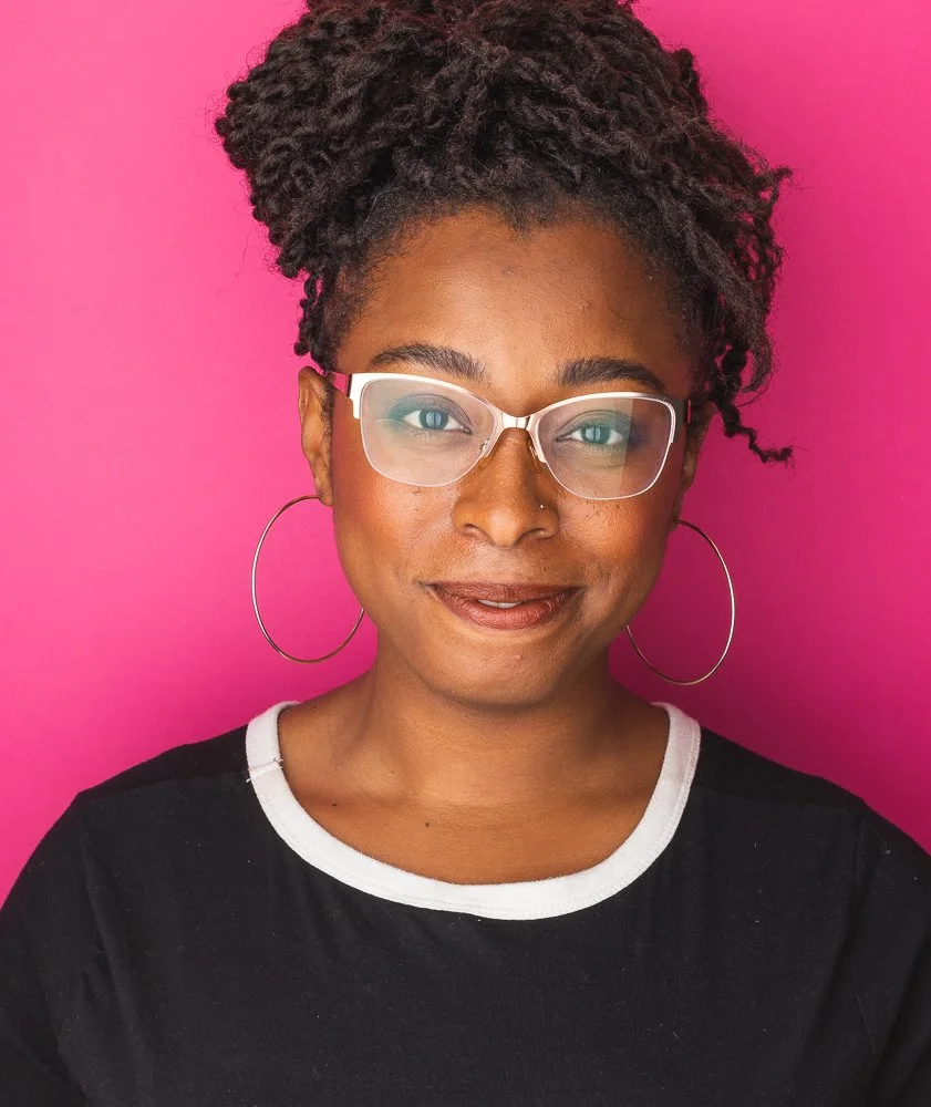 A woman with curly hair styled in a high bun, wearing glasses, large hoop earrings, and a black shirt with a white neckline, smiling against a pink background.