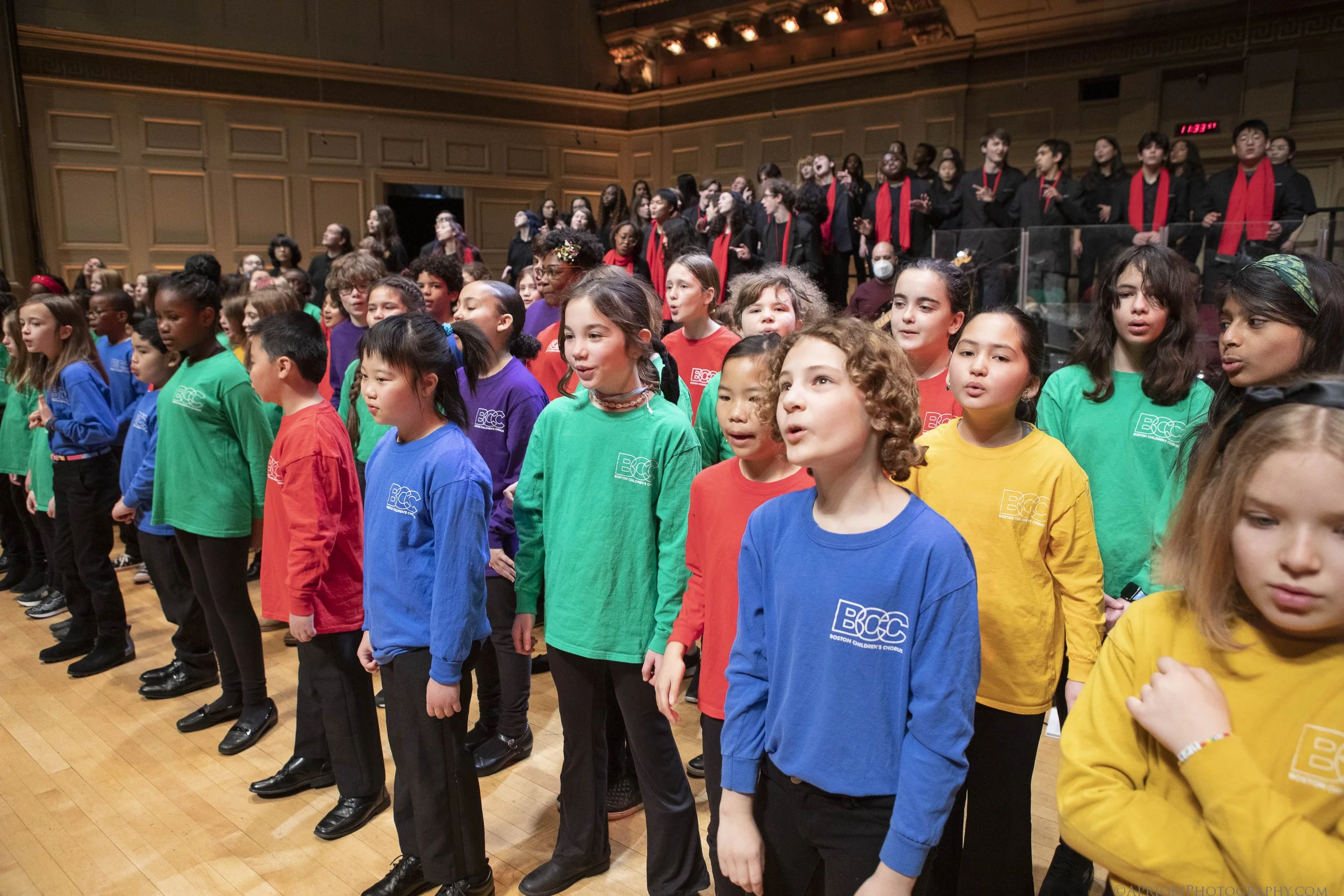 Children wearing colorful shirts standing in a line in a concert hall with a choir singing in the background.