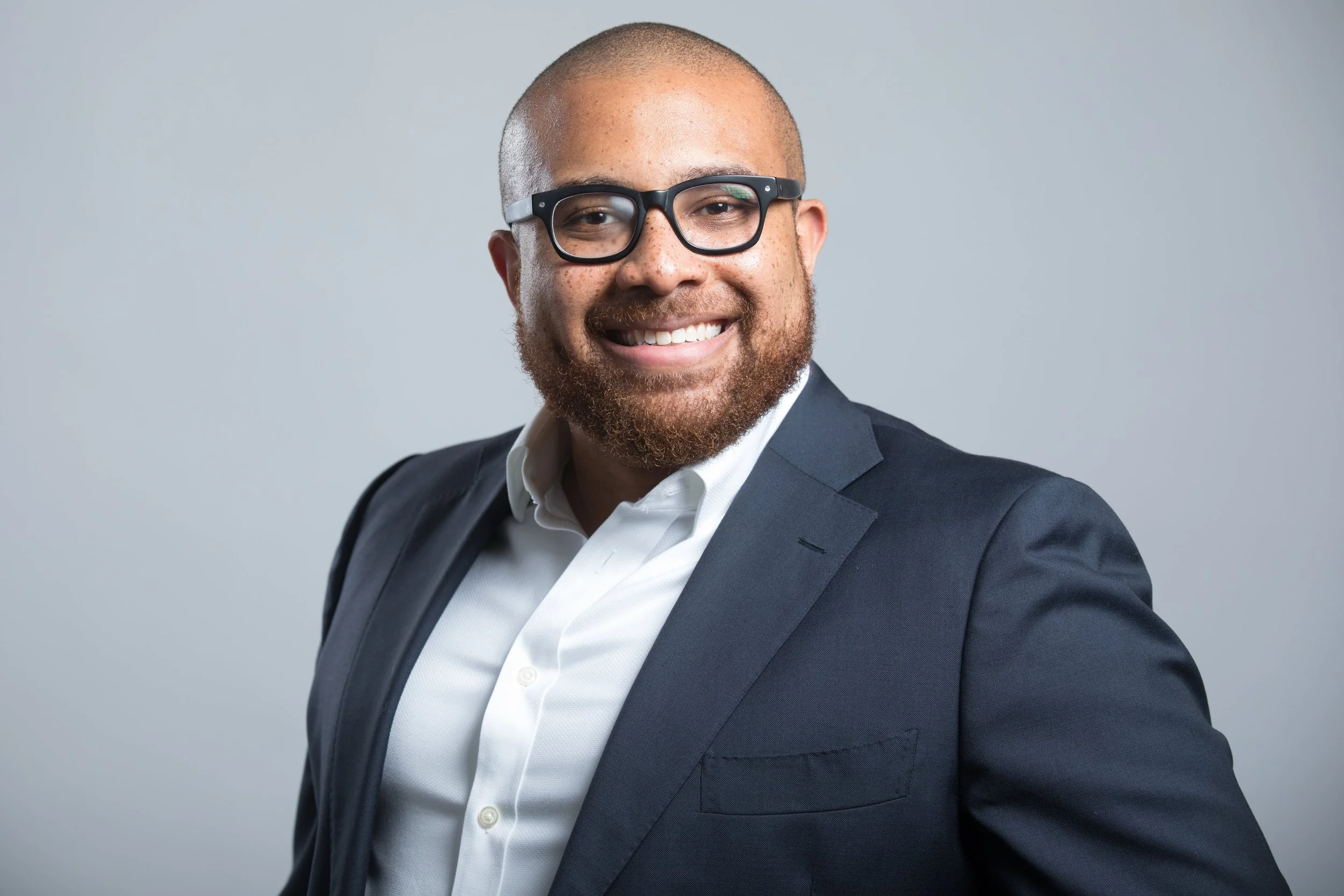 A smiling man with a beard and glasses wearing a dark blazer and white shirt against a light grey background.