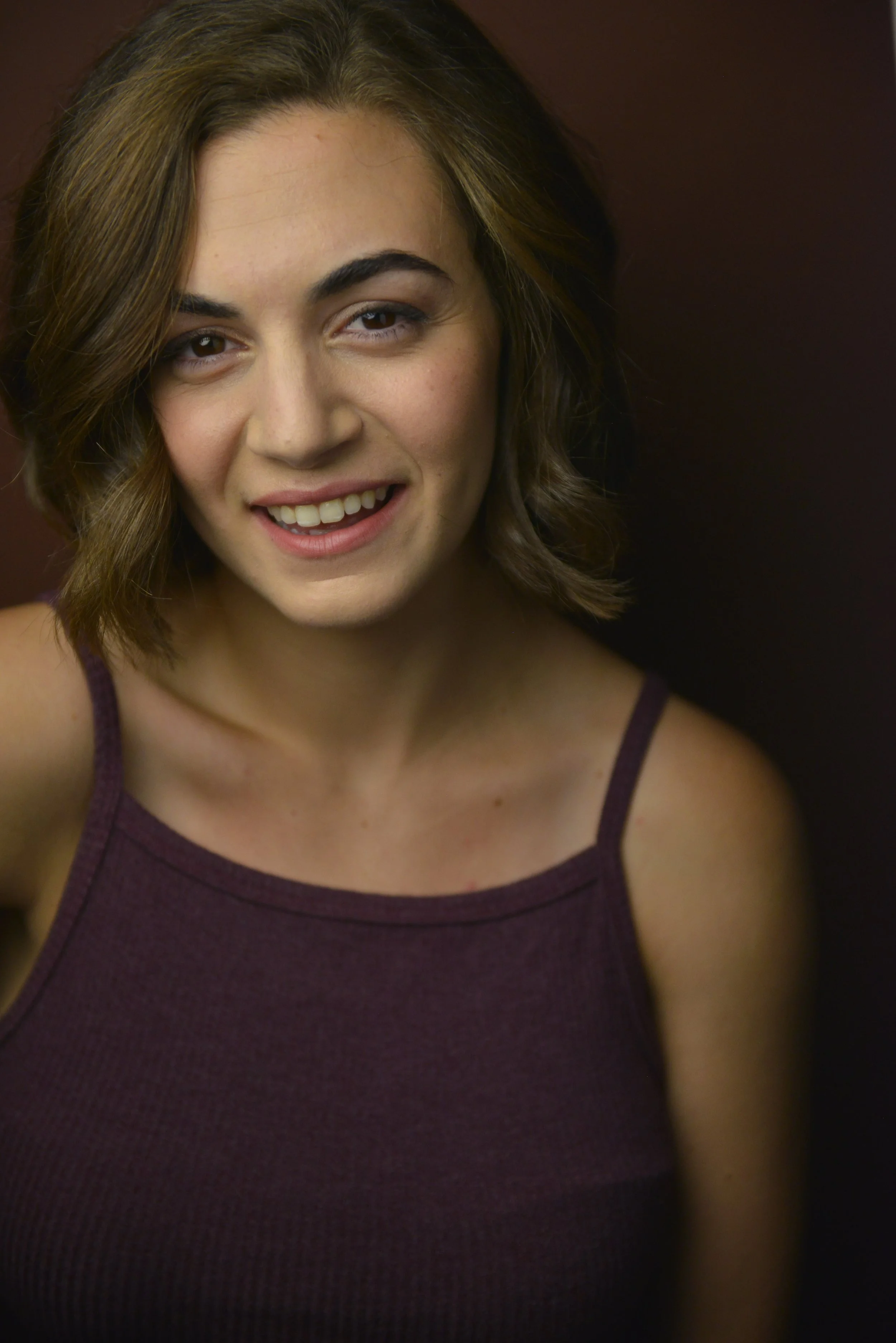 Close-up of a young woman with shoulder-length brown hair, smiling, wearing a sleeveless maroon top, against a dark background.