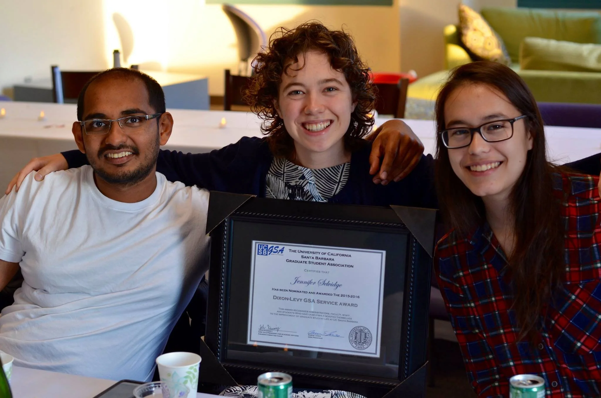 Three people smiling and sitting at a table, one holding a framed certificate of appreciation from the University of California, Santa Barbara, Graduate Student Association, awarded to Jennifer Sevidge for service during 2015-2016.