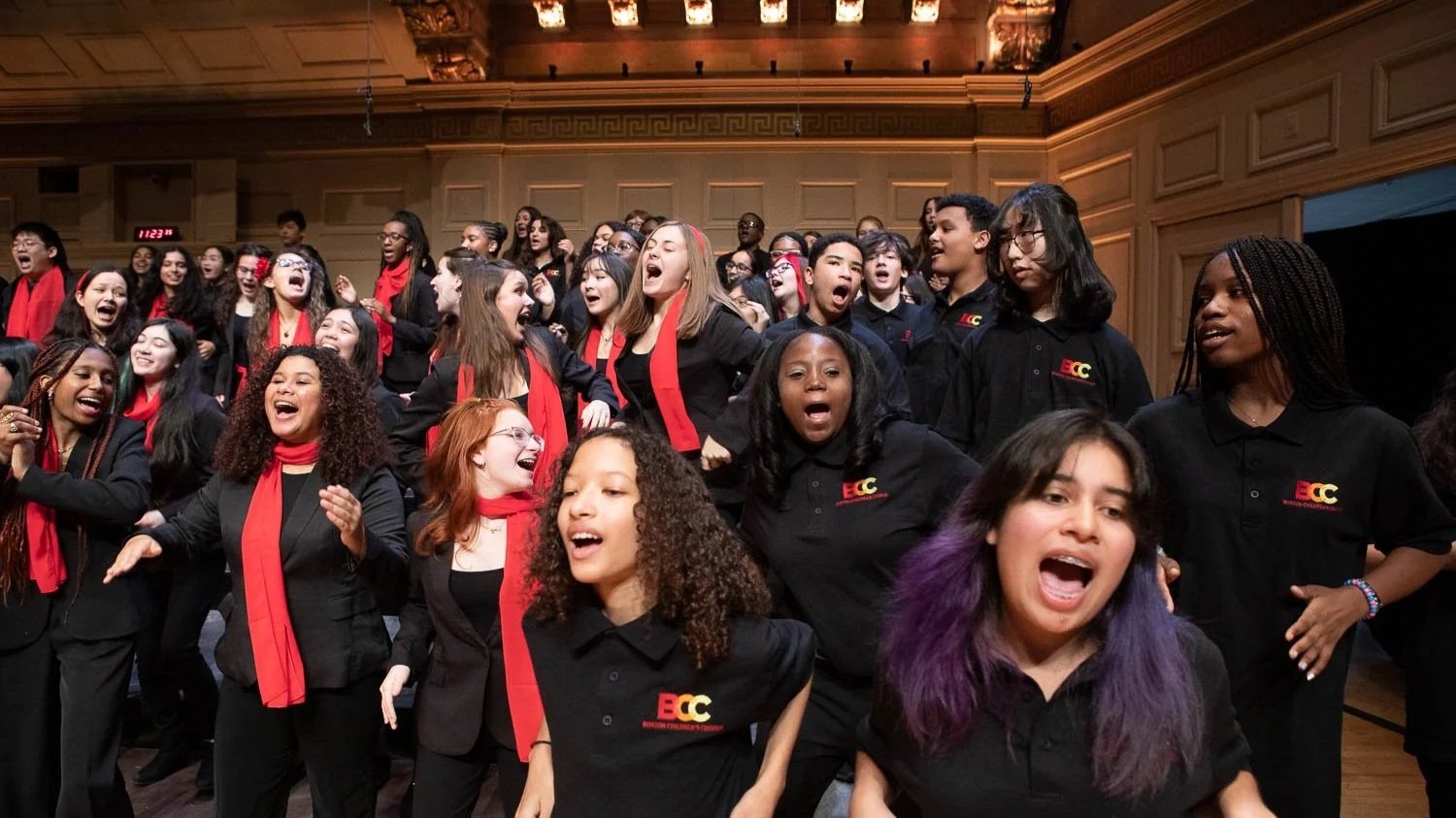 Group of children and teenagers singing passionately on stage during a choir concert at a formal auditorium, many wearing black outfits with red scarves or accessories.